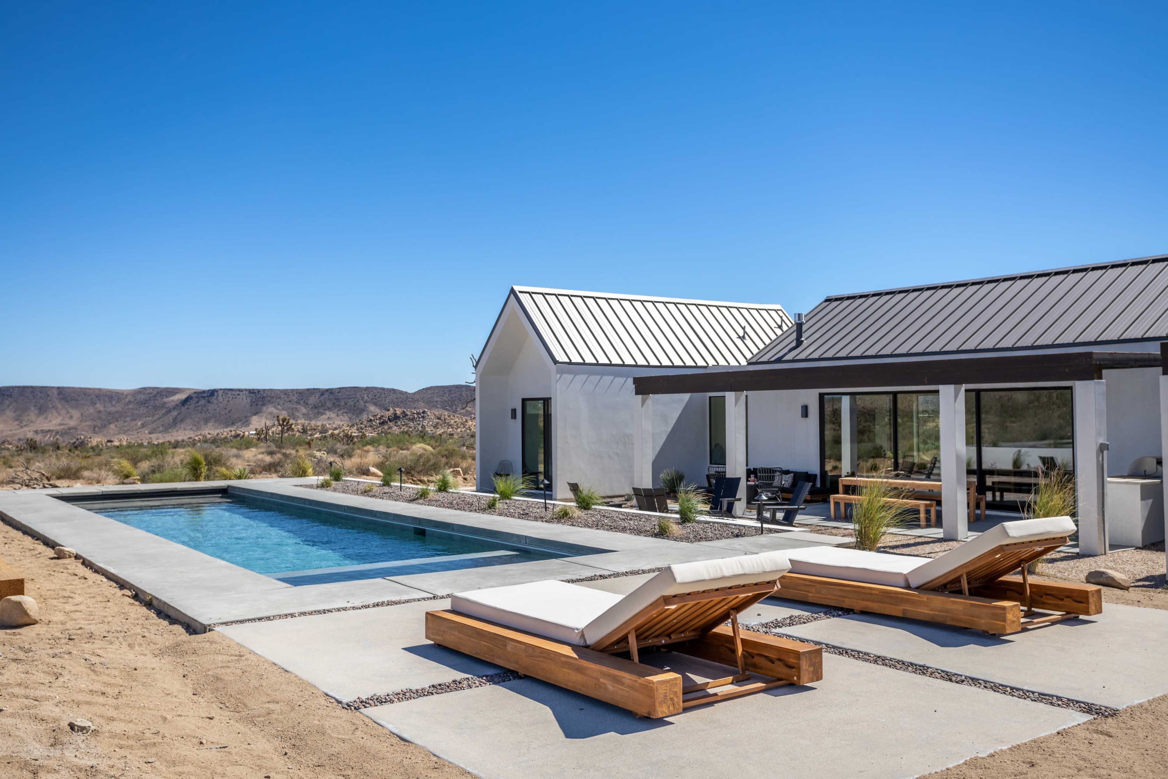 A modern house with a pool and two lounge chairs is set in a desert landscape under a clear blue sky.