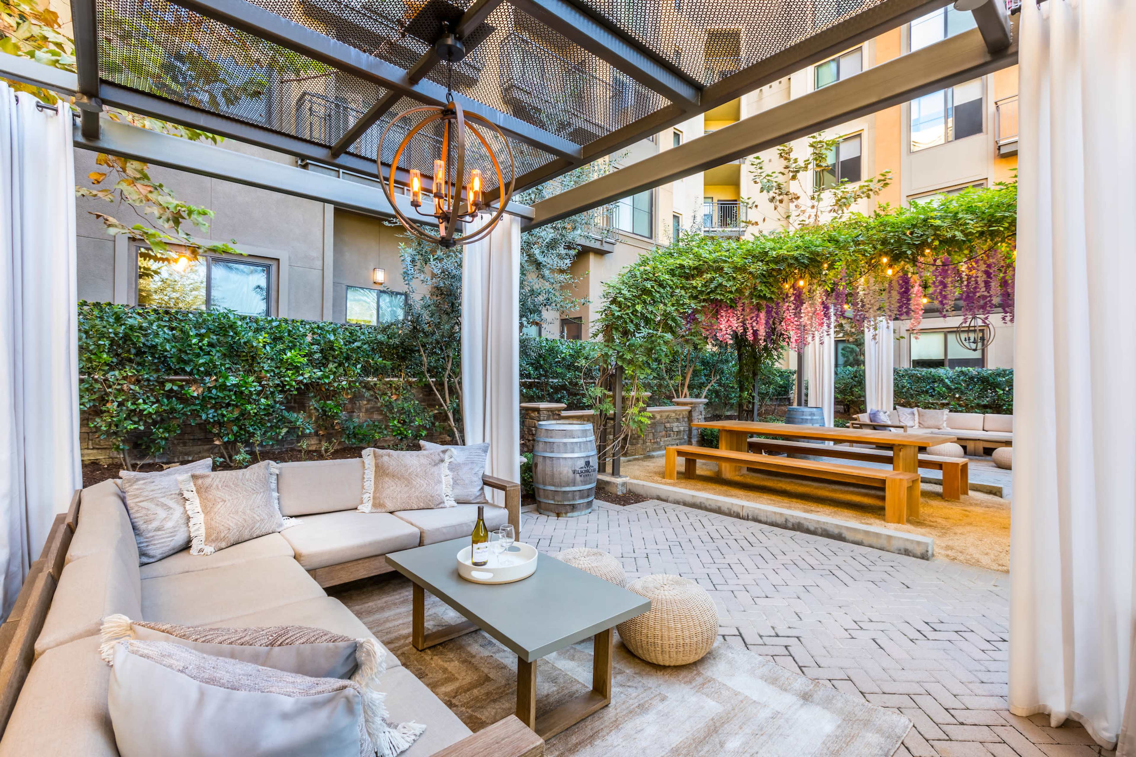 A landscaped outdoor seating area with a sofa, a coffee table, and a dining table surrounded by greenery and decorative lighting.