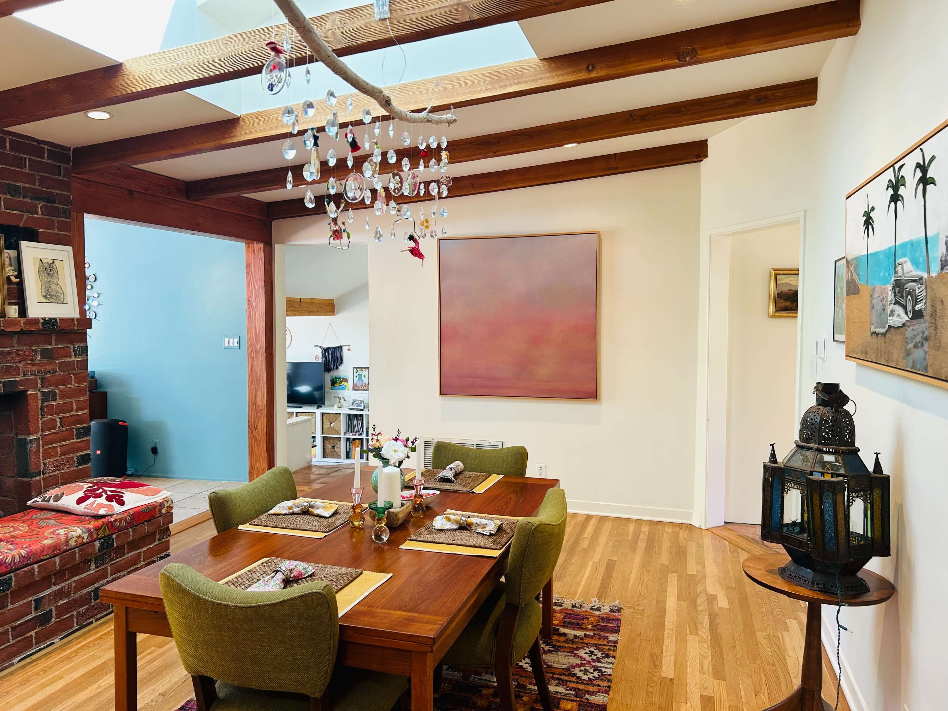 A dining area features a wooden table set for a meal, surrounded by green chairs, with a chandelier hanging above and artwork on the walls.