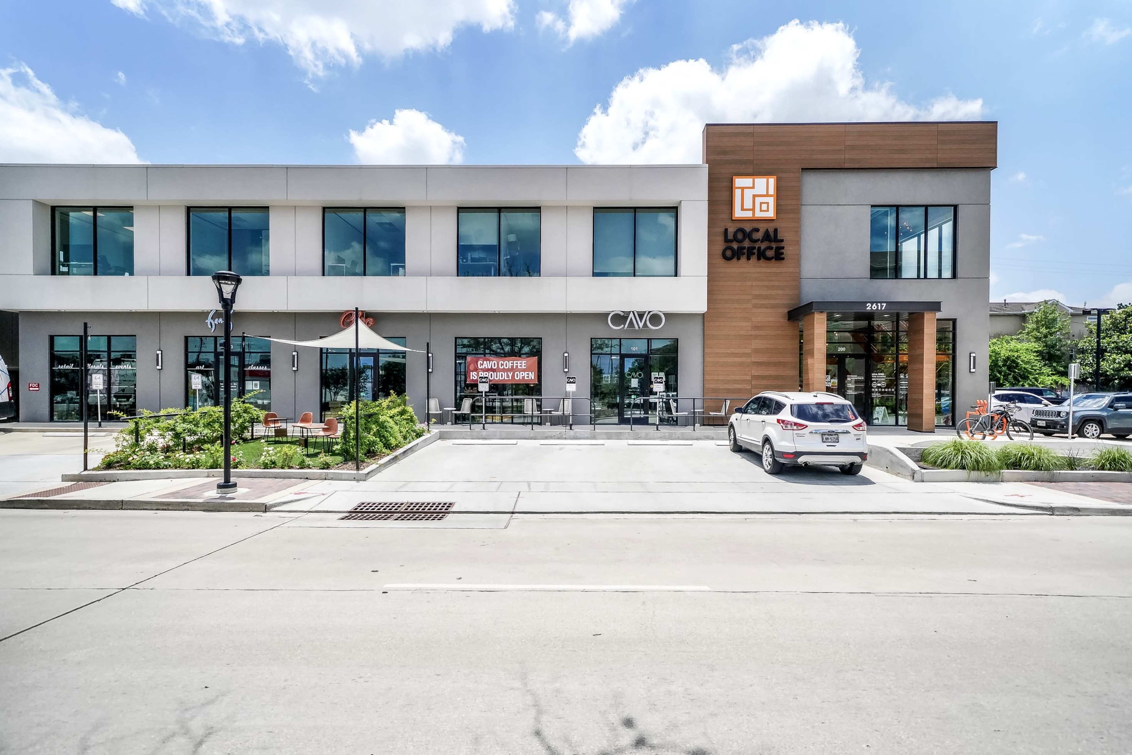 The image shows a modern commercial building with a restaurant and office space, featuring outdoor seating and parked cars along a paved street.