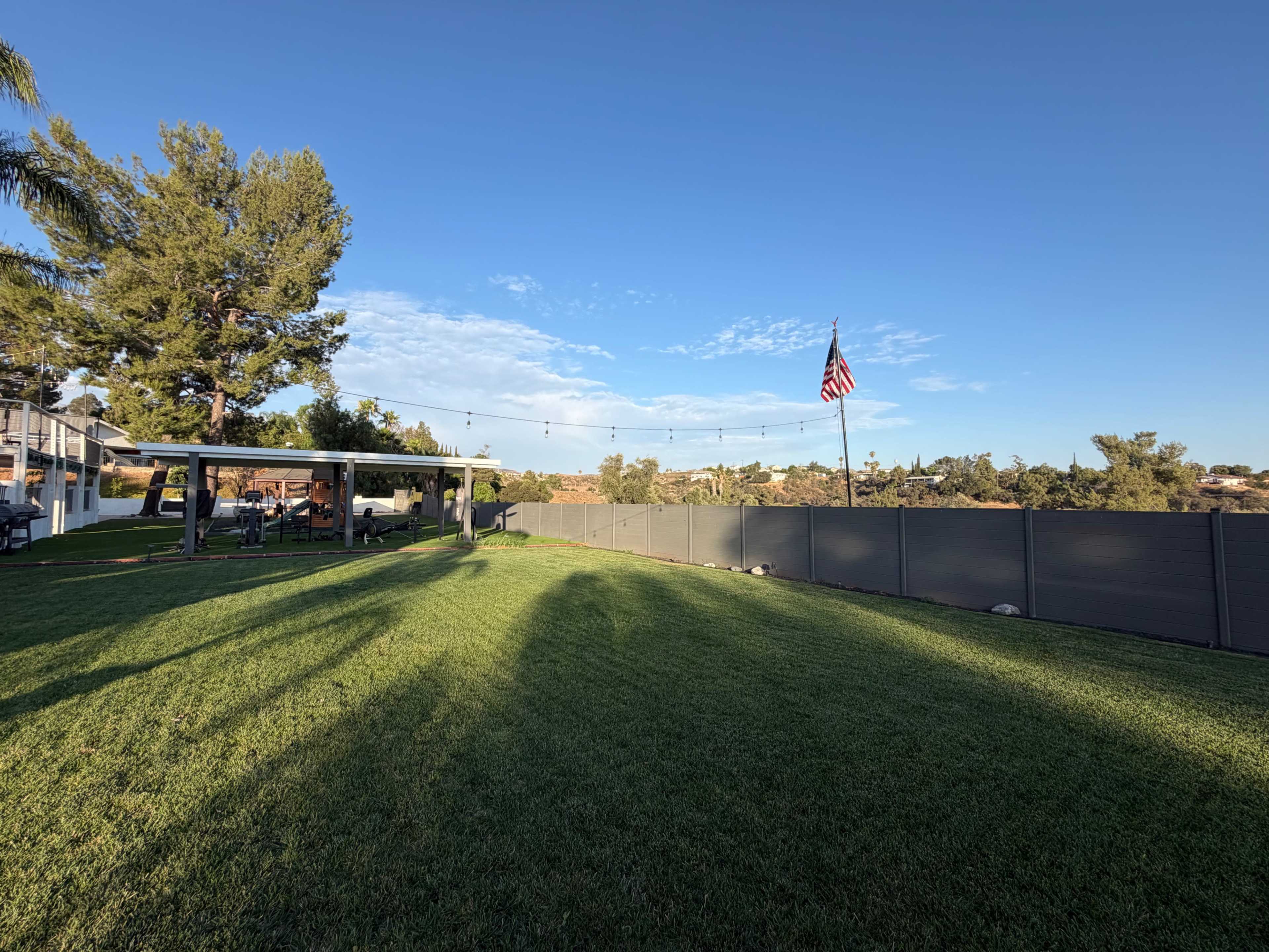 The image depicts a grassy backyard with a pergola, a wooden deck, string lights, and a flagpole with an American flag, all set against a clear blue sky.