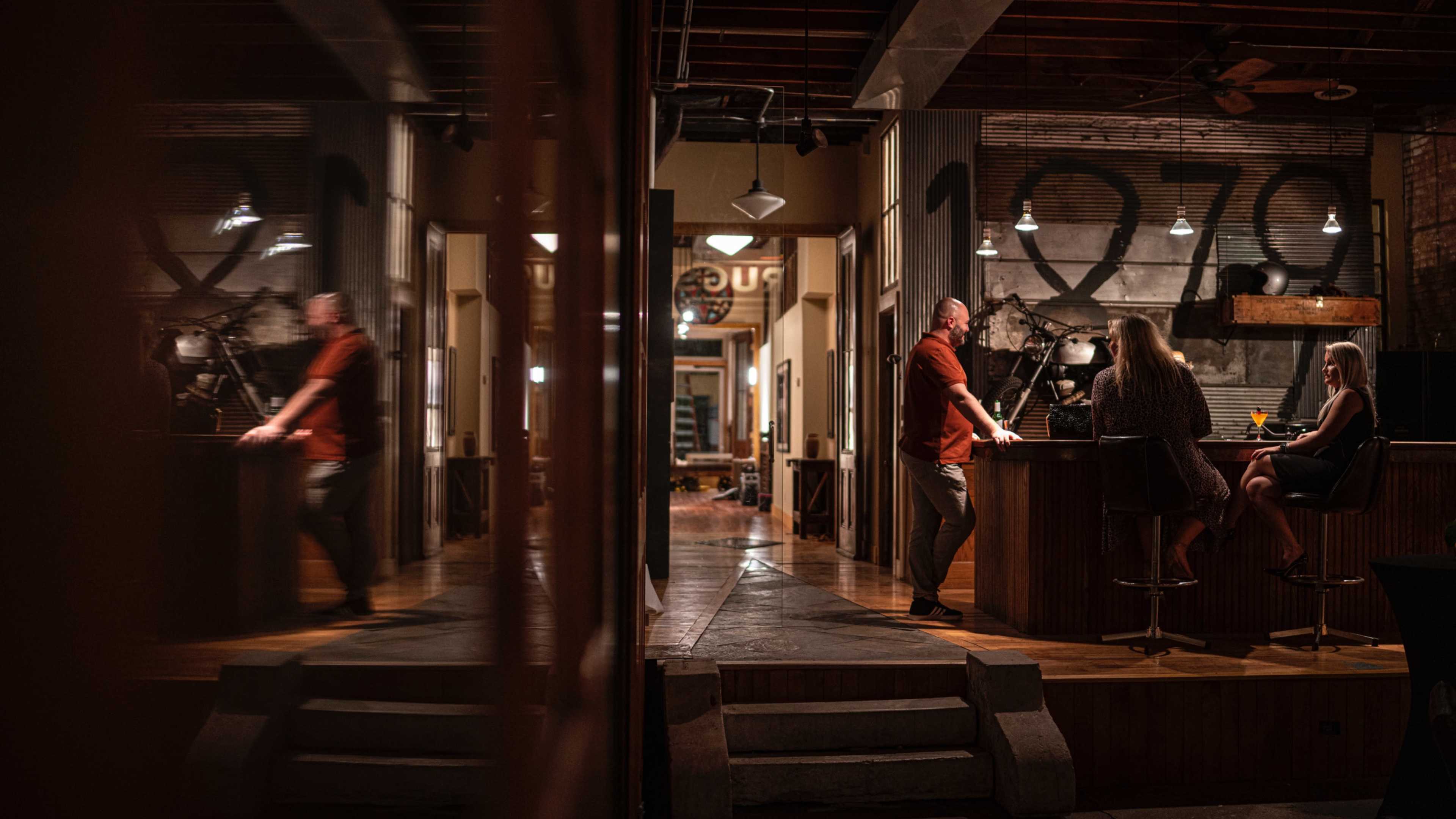 A bar scene features two patrons sitting at the counter while a server stands nearby in a dimly lit establishment with industrial decor.