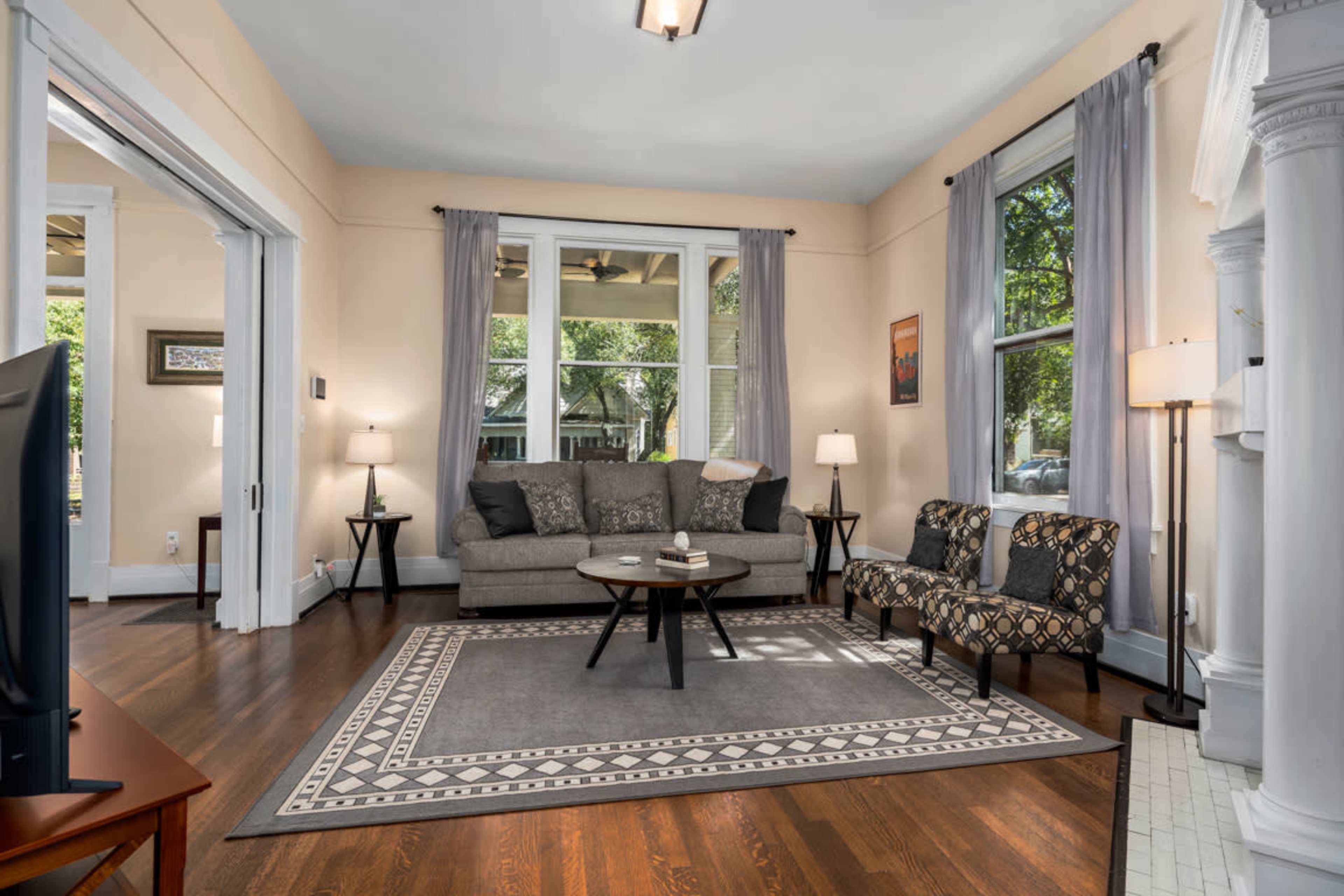 A living room with a gray sofa, a patterned chair, a coffee table, and large windows allowing natural light.