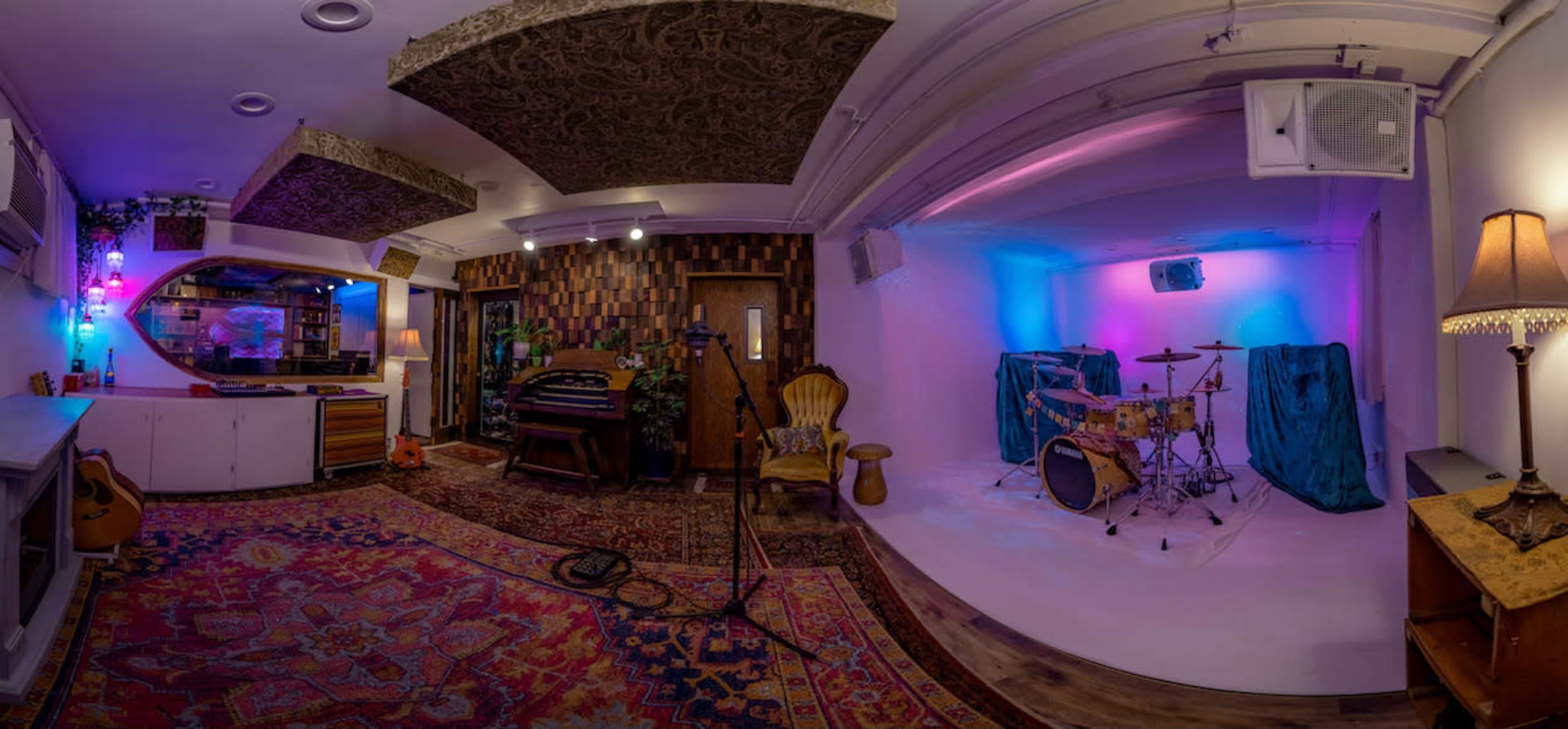A music studio featuring a drum set, colorful lighting, a patterned rug, and various musical instruments along with decorative plants.