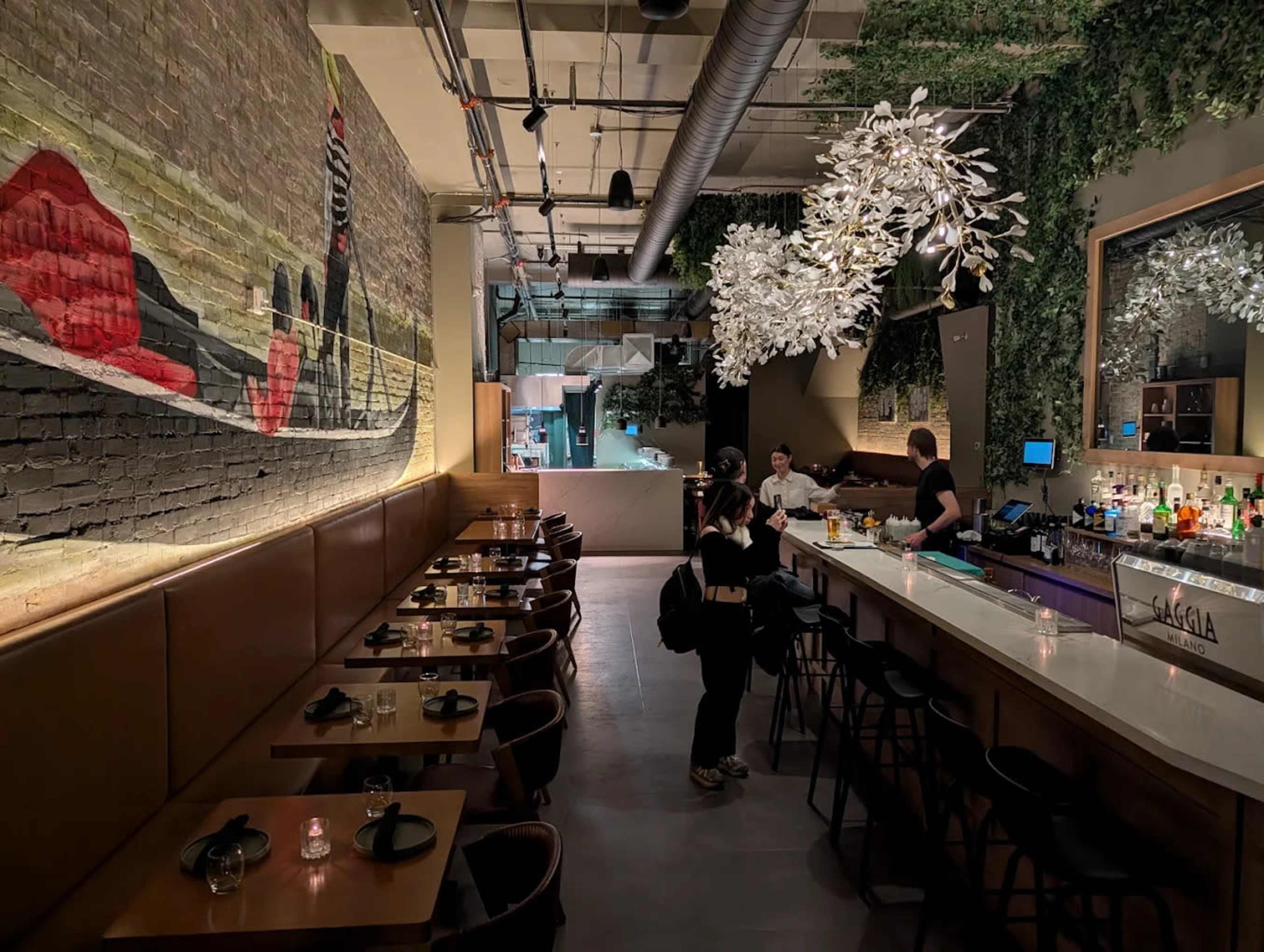 The image shows a modern bar area with a long counter, decorative wall art, and tables arranged for dining, surrounded by greenery and stylish lighting.