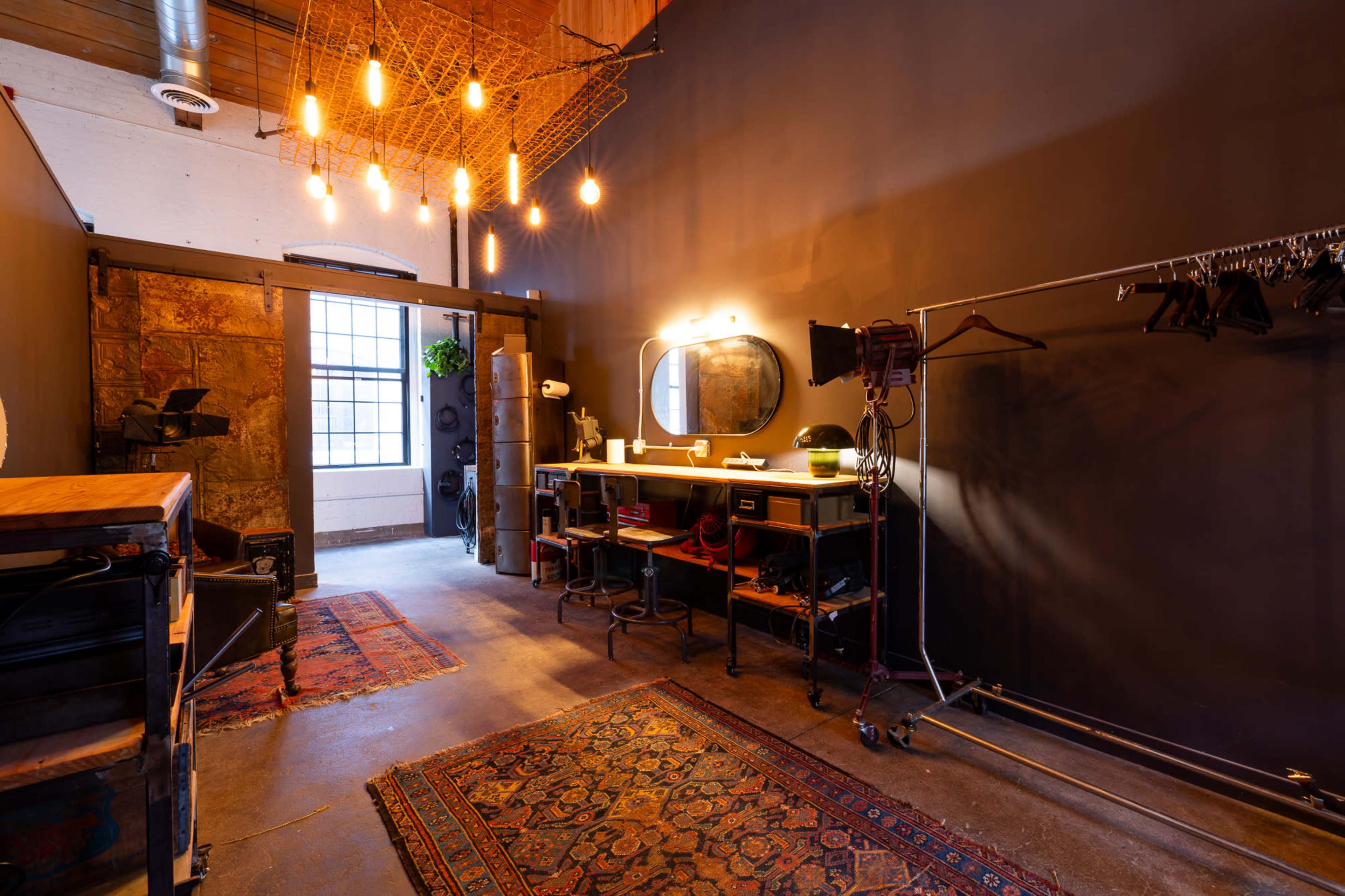 A dimly lit room with a dark wall, featuring a mirror, a stylish vanity setup, and a clothing rack, all illuminated by hanging light bulbs.