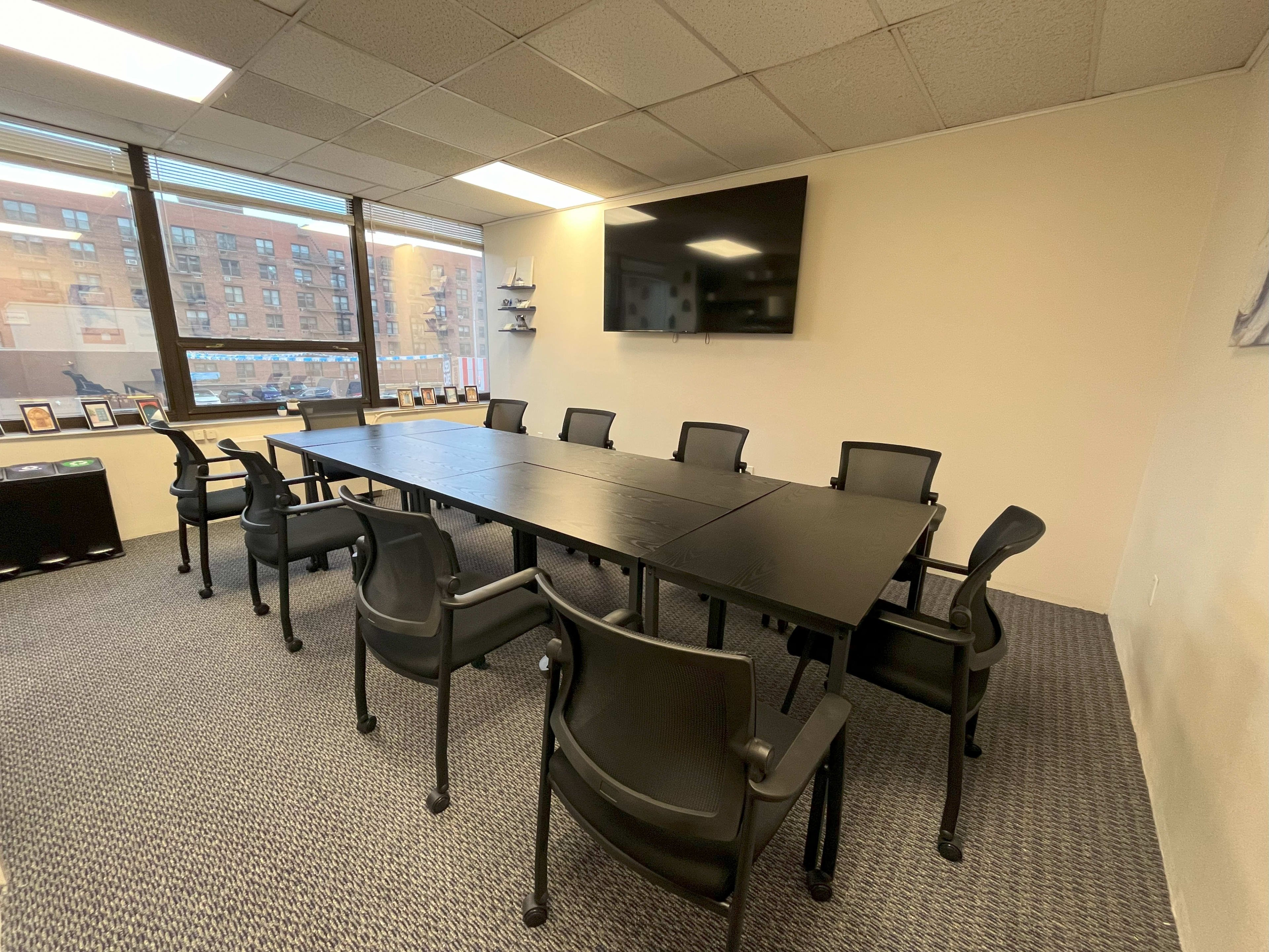 A conference room with a long black table surrounded by ten black chairs and a wall-mounted television.