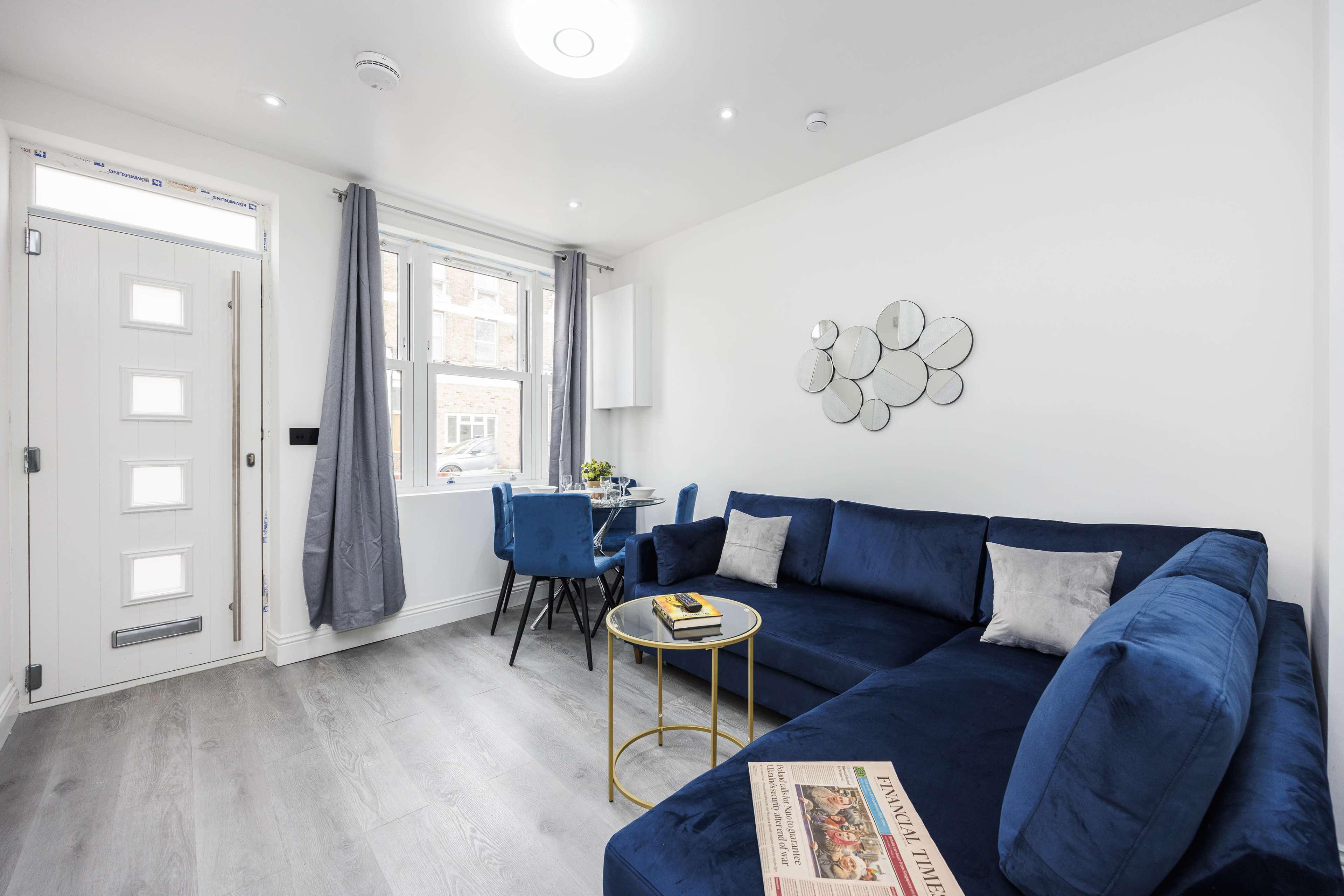 The image shows a modern living room with a blue sectional sofa, a round coffee table, and a door leading outside, complemented by a dining area visible in the background.