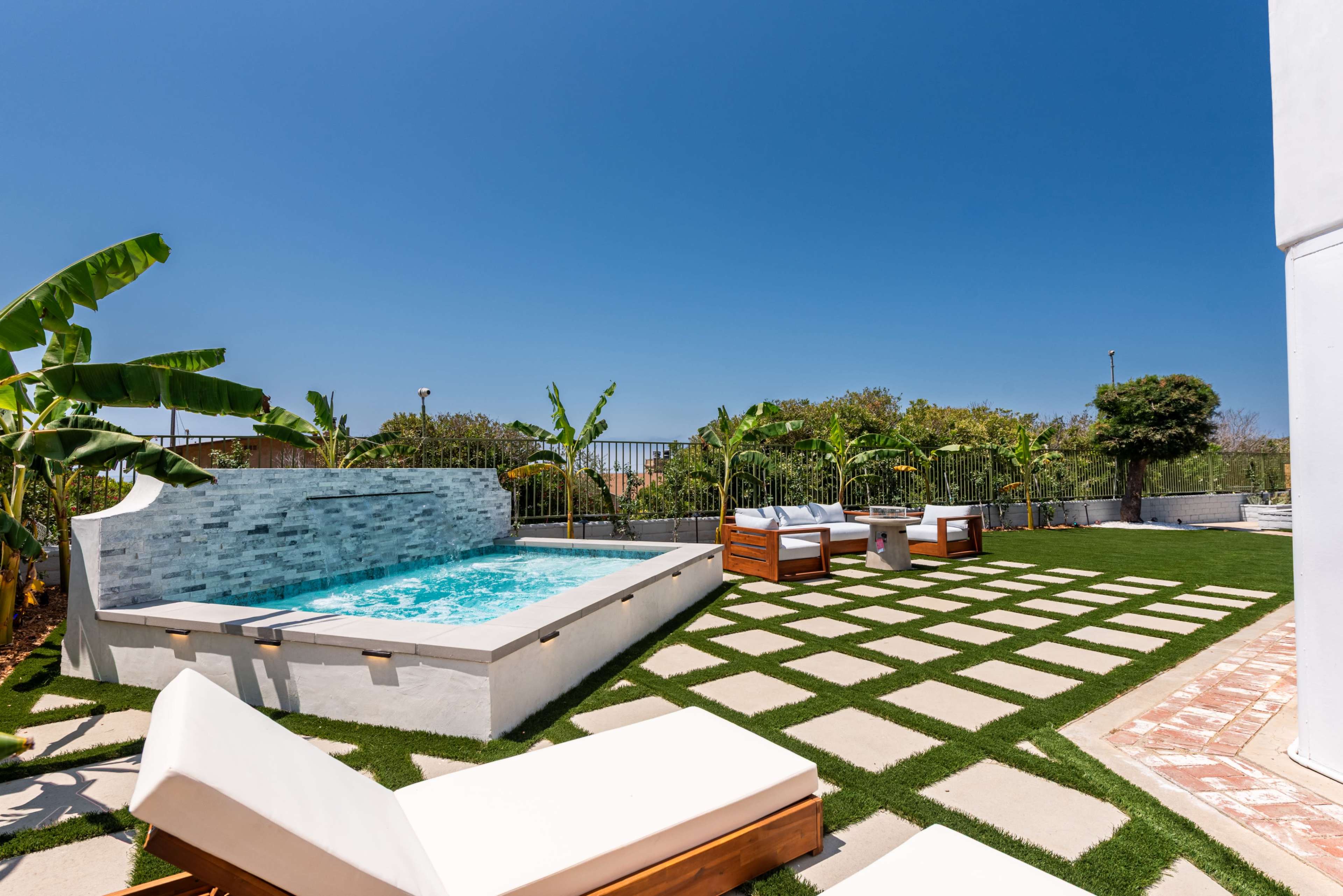 A rectangular hot tub is located on a grassy area surrounded by banana trees and patio furniture under a clear blue sky.