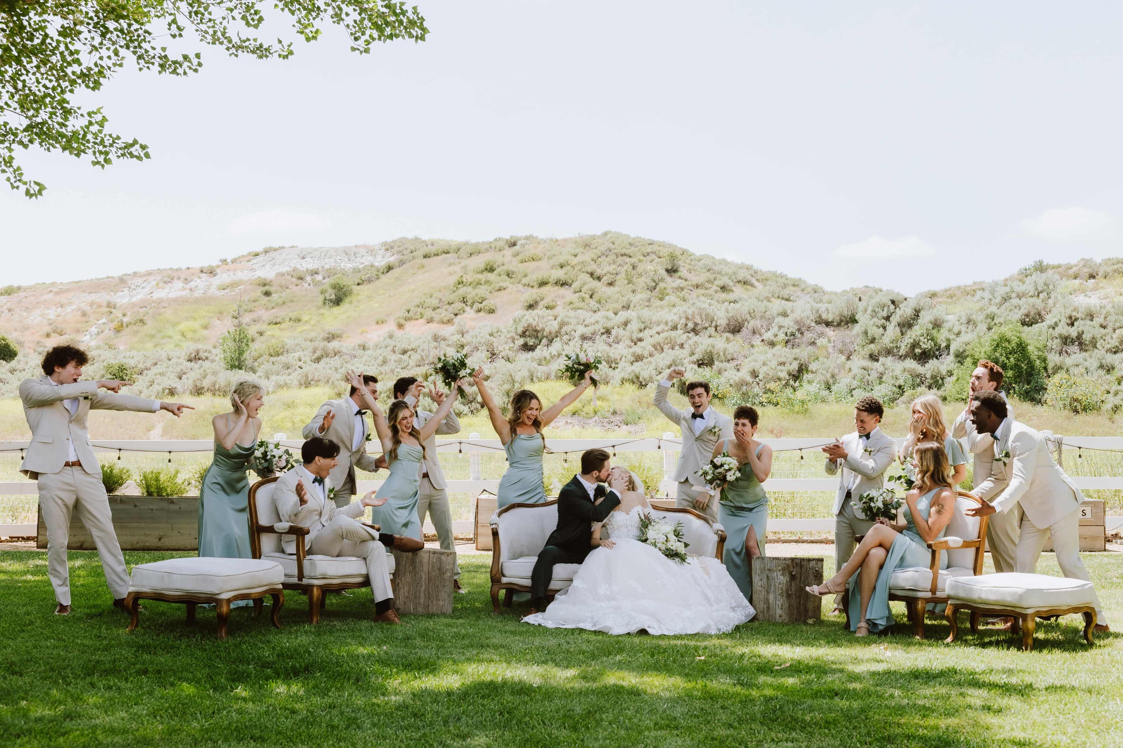 A wedding party poses outdoors, with the bride and groom seated in front while friends and family strike playful poses around them.