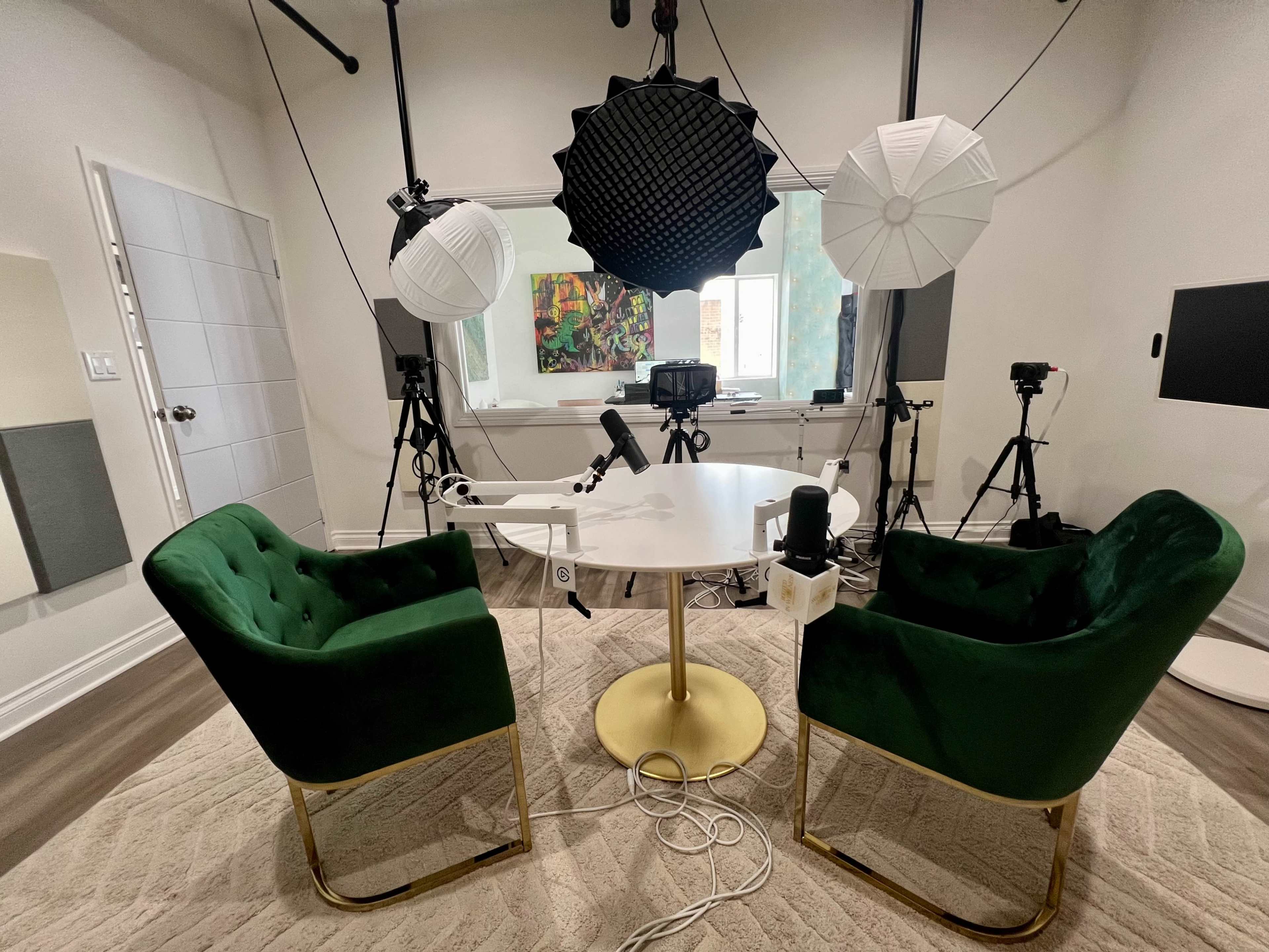 A modern recording studio features two green chairs positioned around a circular table, with studio lights, cameras, and sound equipment arranged in the background.