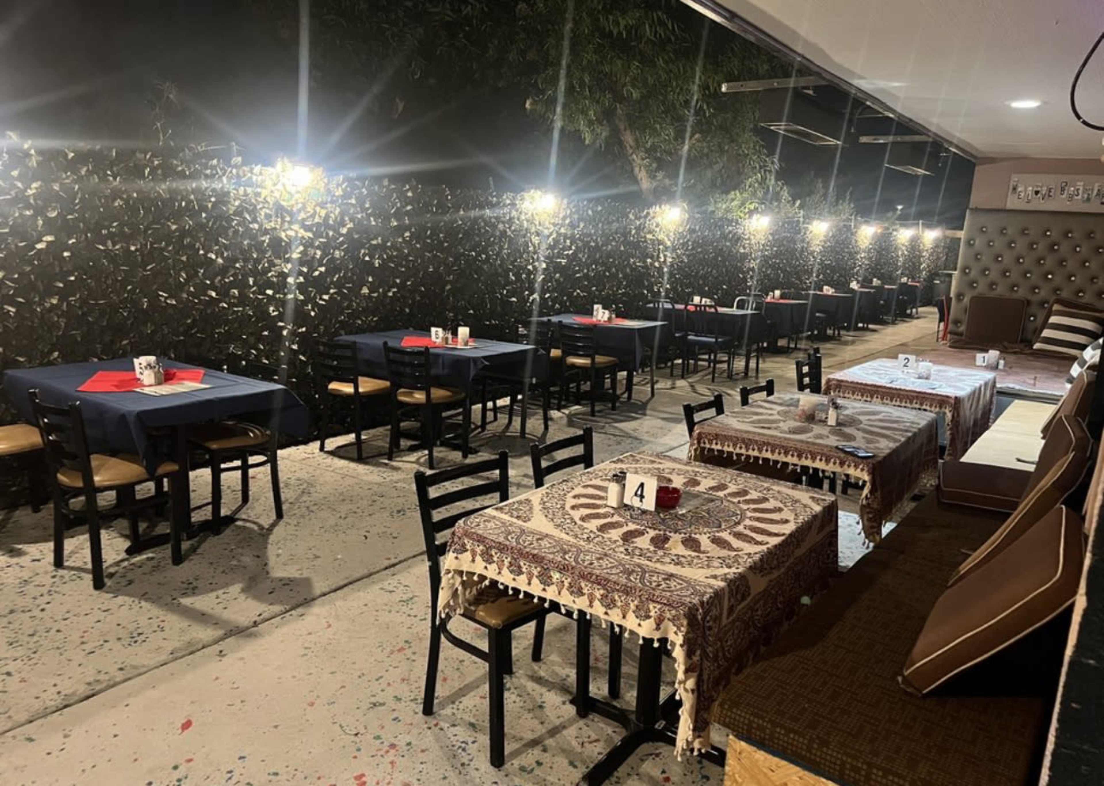 A dimly lit outdoor dining area with several tables set for customers, featuring tables covered with tablecloths and decorative runners.
