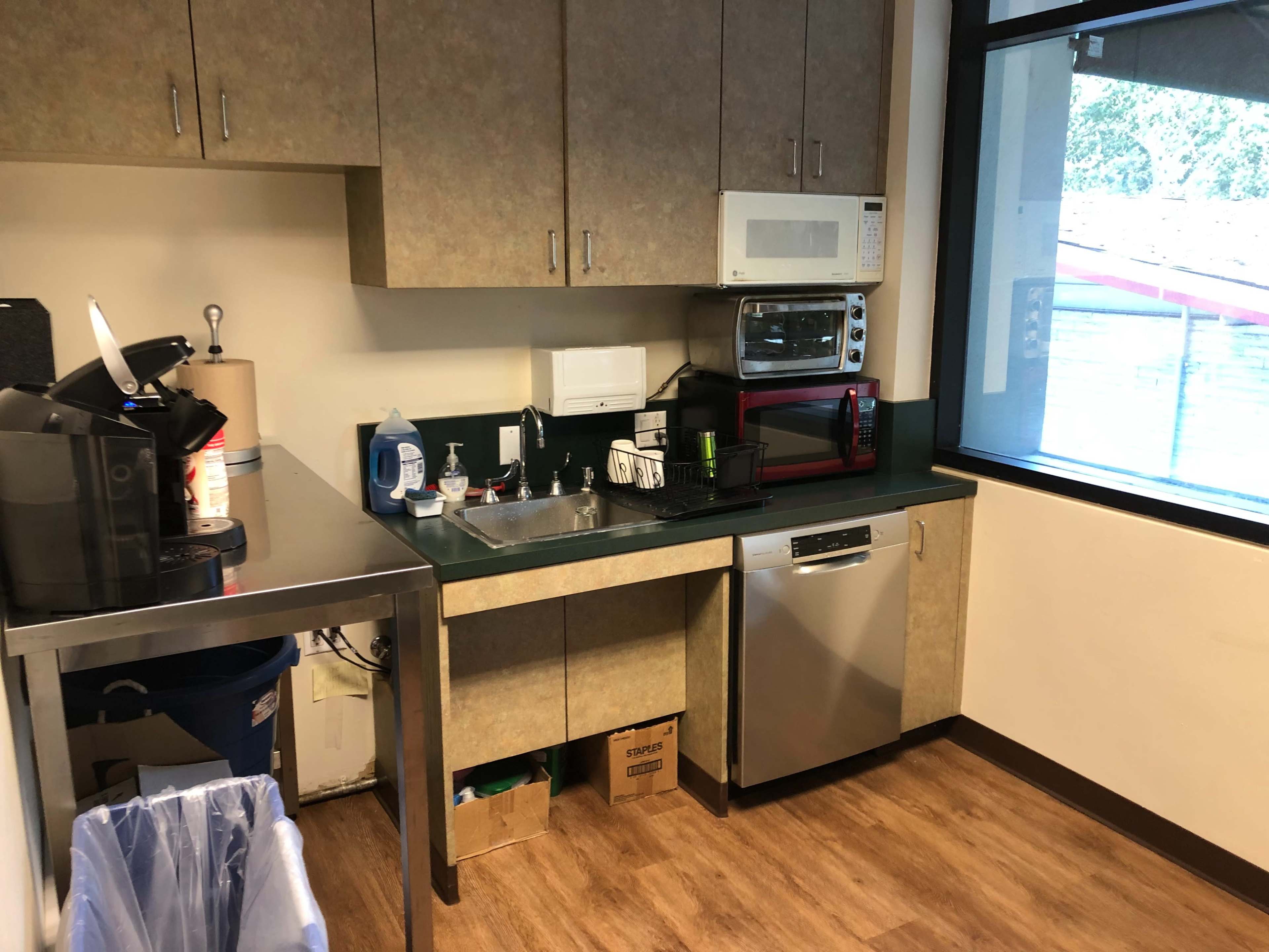 The image shows a kitchen area with a sink, microwave, coffee maker, and various appliances on the counter, along with a trash bin under the counter.
