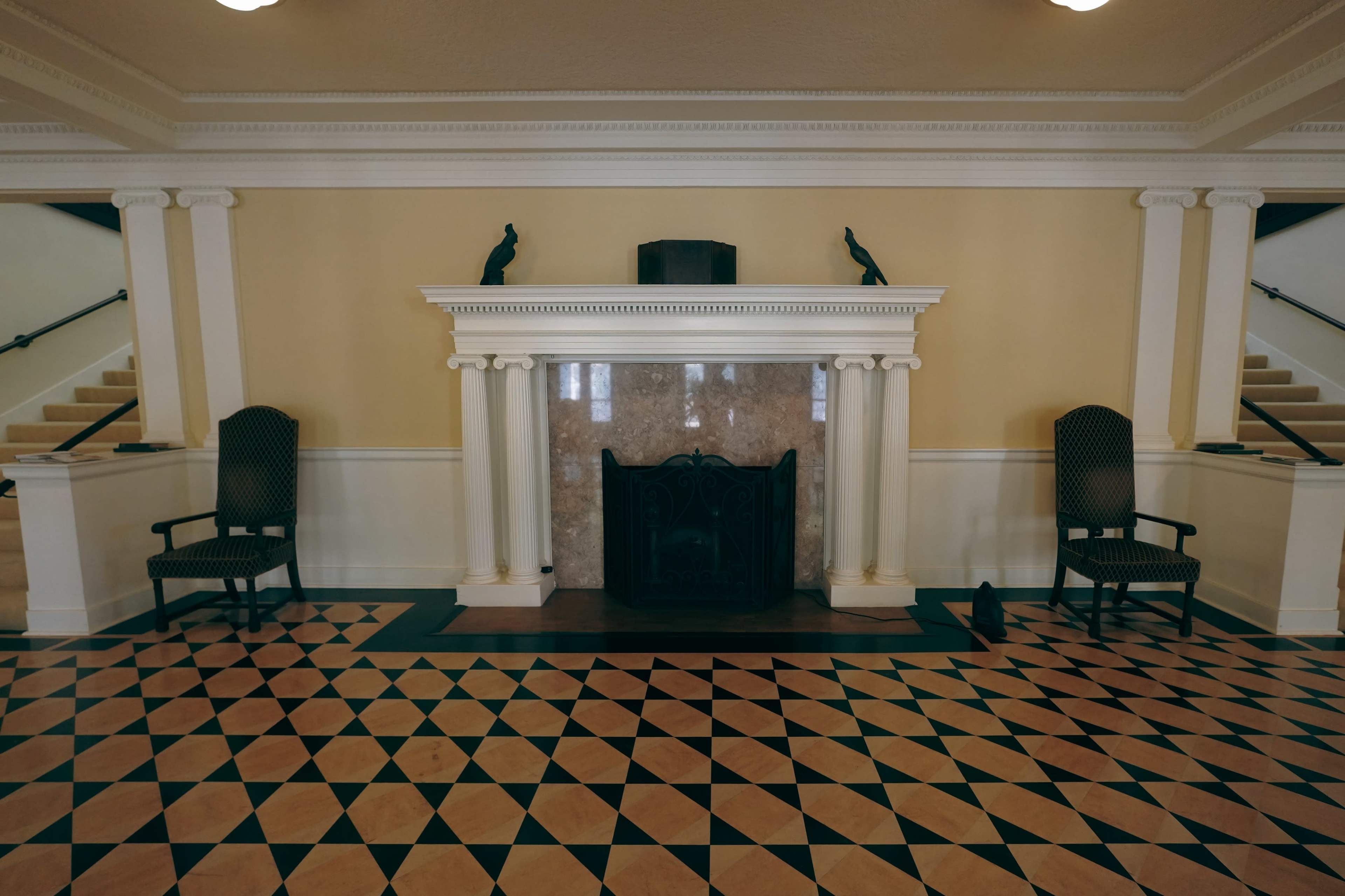 An interior space with a marble fireplace centered against a pale wall, flanked by two black chairs, and a patterned tile floor in shades of blue and beige.