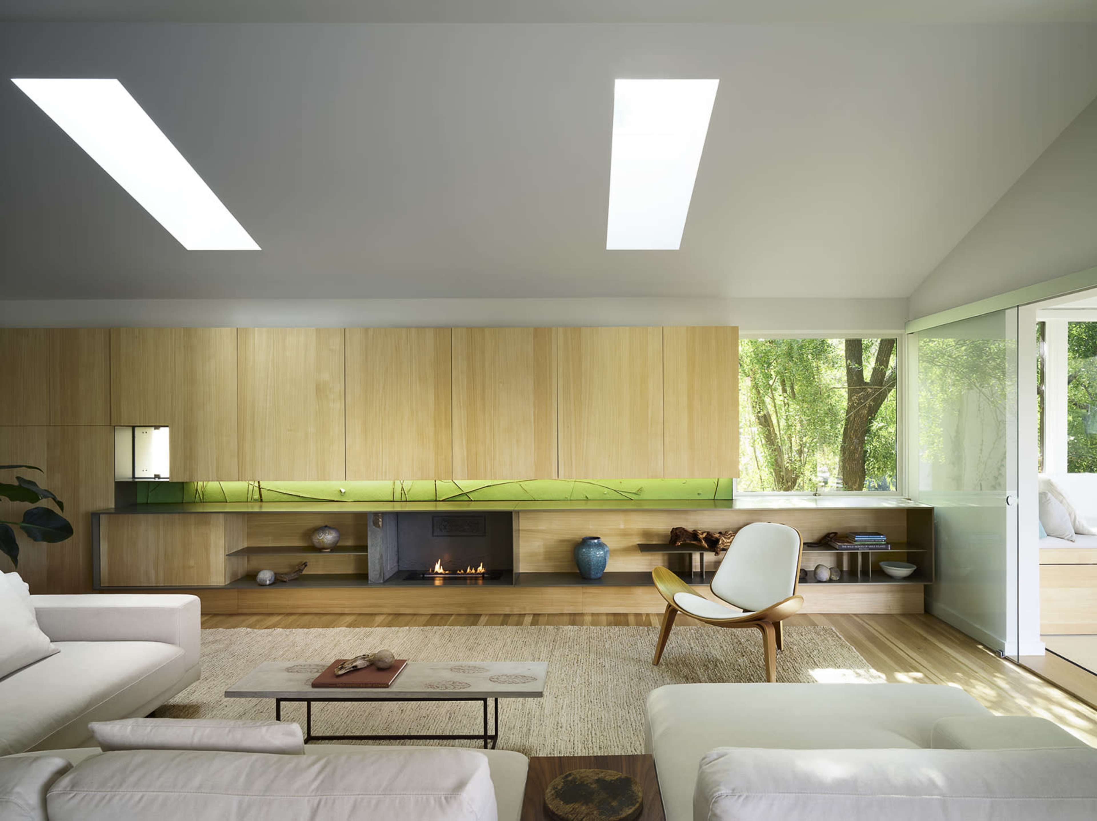 The image shows a modern living room with light wood cabinetry, a fireplace, and large windows that bring in natural light.