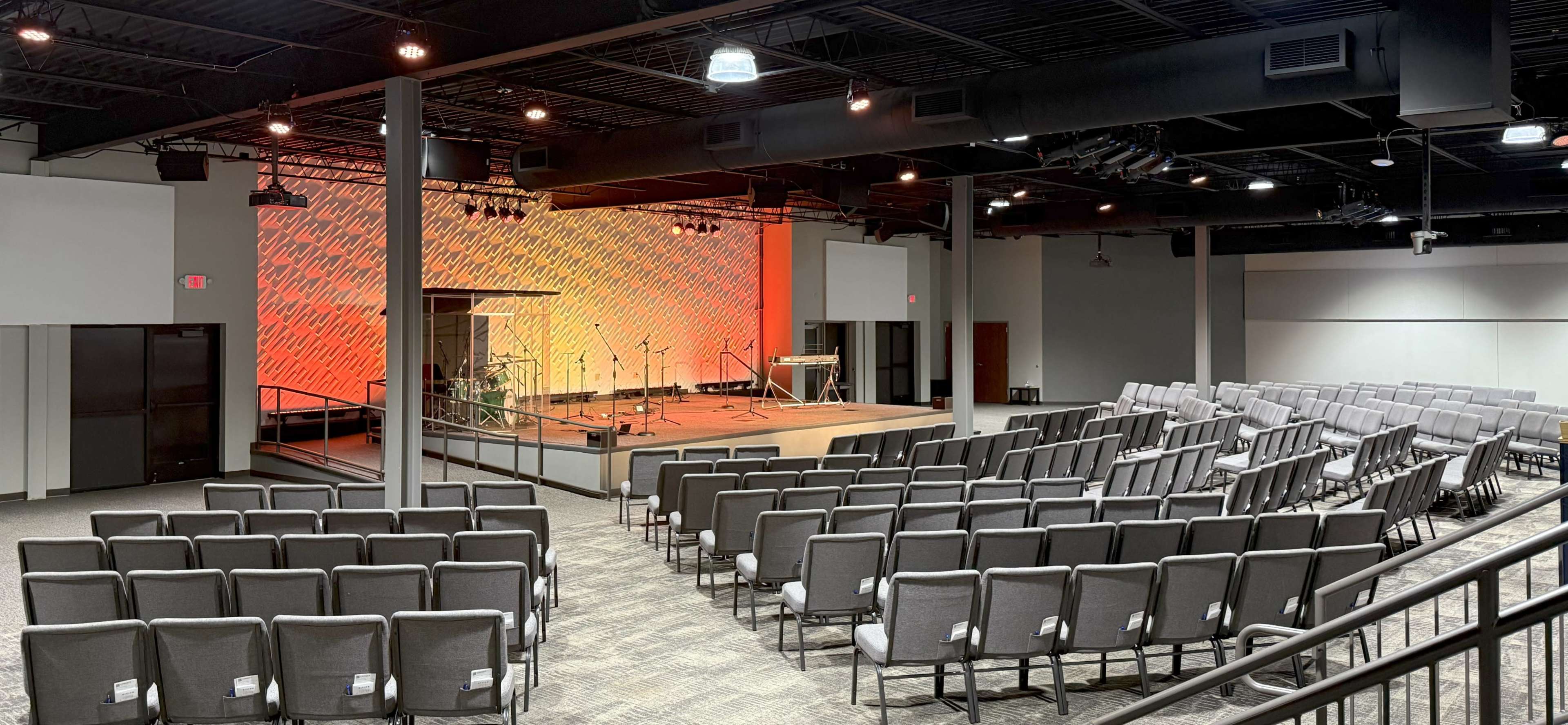 The image shows an interior of a modern auditorium with rows of gray chairs facing a stage, illuminated by a colorful backdrop and equipped with musical instruments.