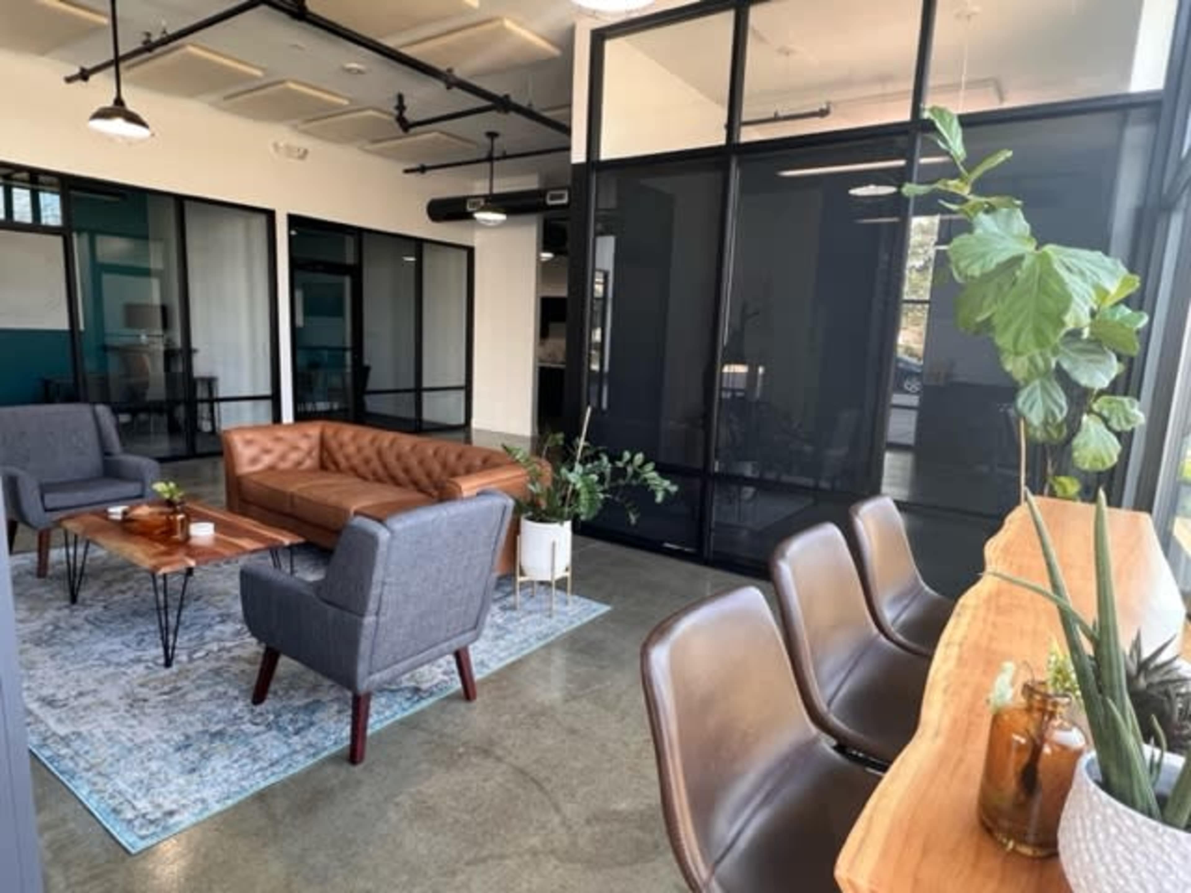 A modern office lounge area with a brown leather sofa, gray armchairs, a wooden table, and large glass partitions.