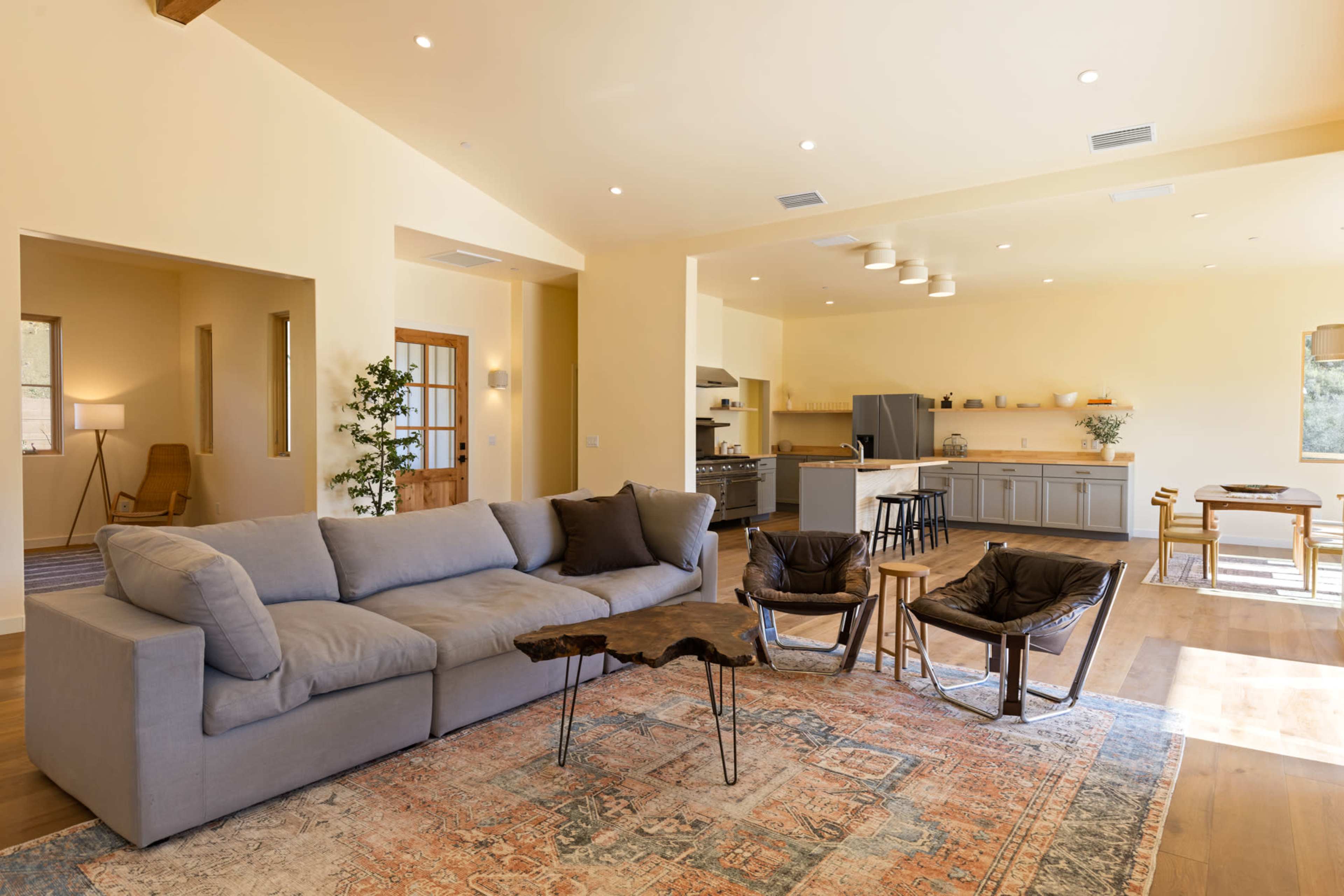 A modern living space with a gray sectional sofa, wooden coffee table, and two armchairs, leading to a kitchen area with gray cabinets and a dining table.