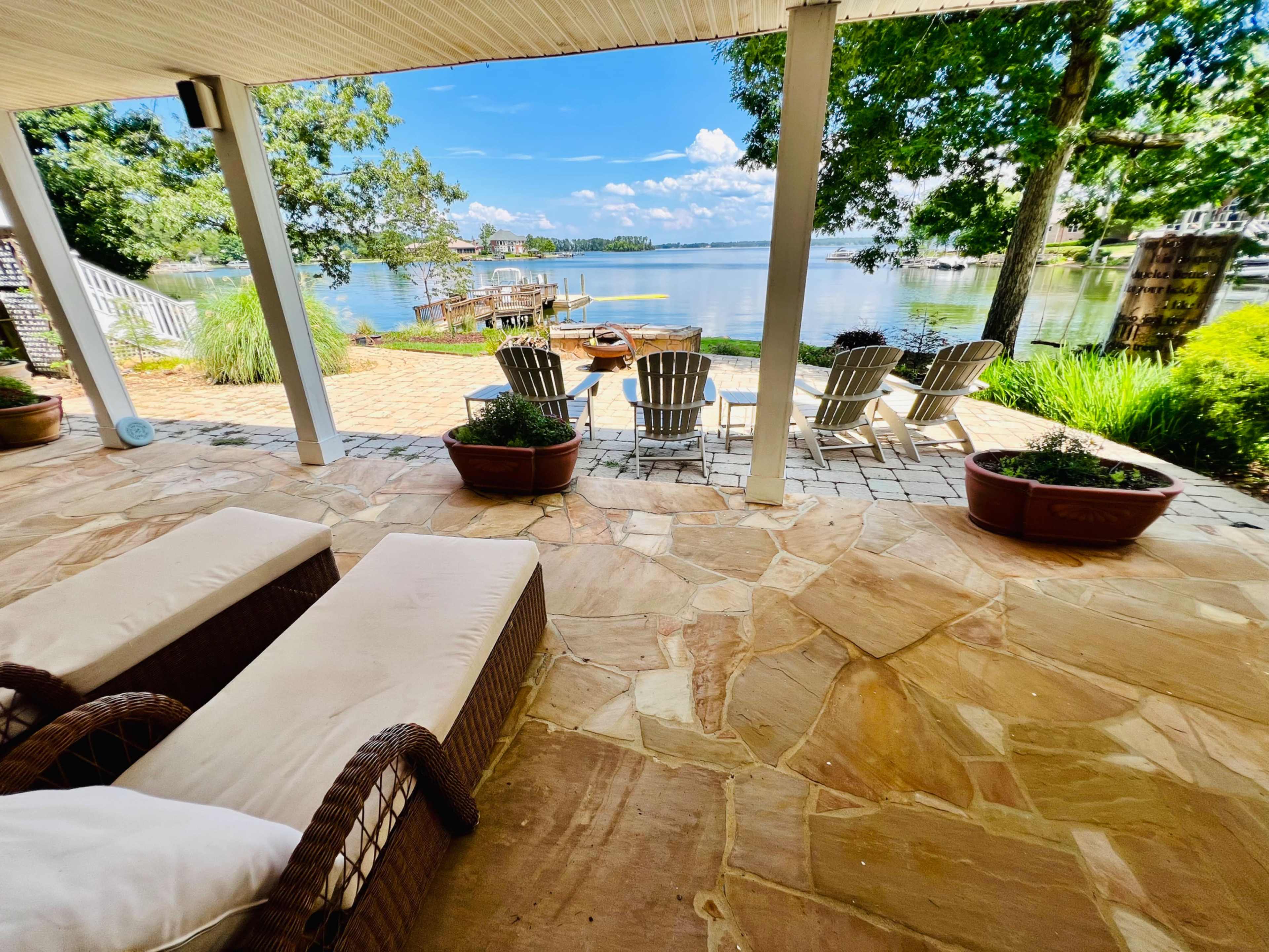 A patio features two lounge chairs overlooking a calm lake, with a dock and several chairs arranged beside the water.