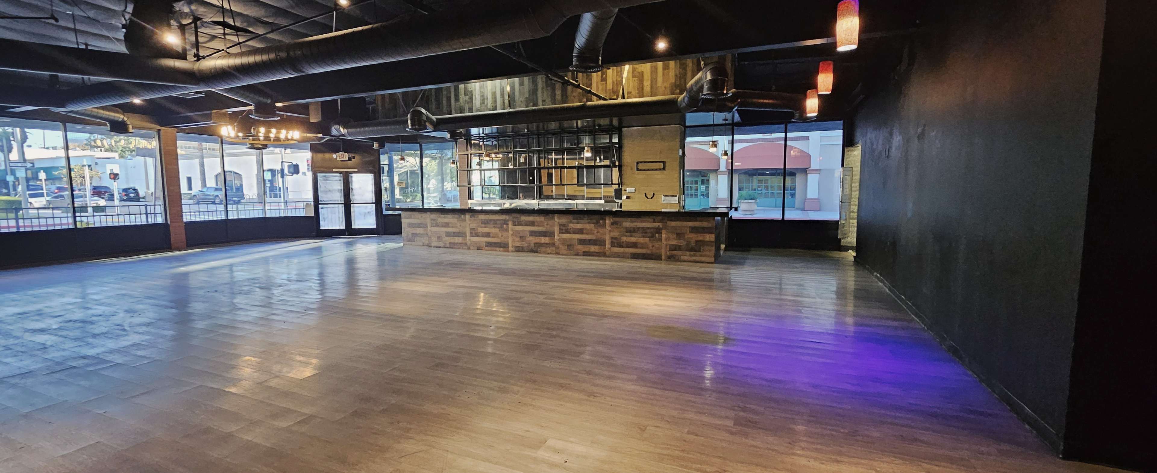 The image shows an empty interior space featuring a wooden bar counter, large windows, and polished wooden flooring.