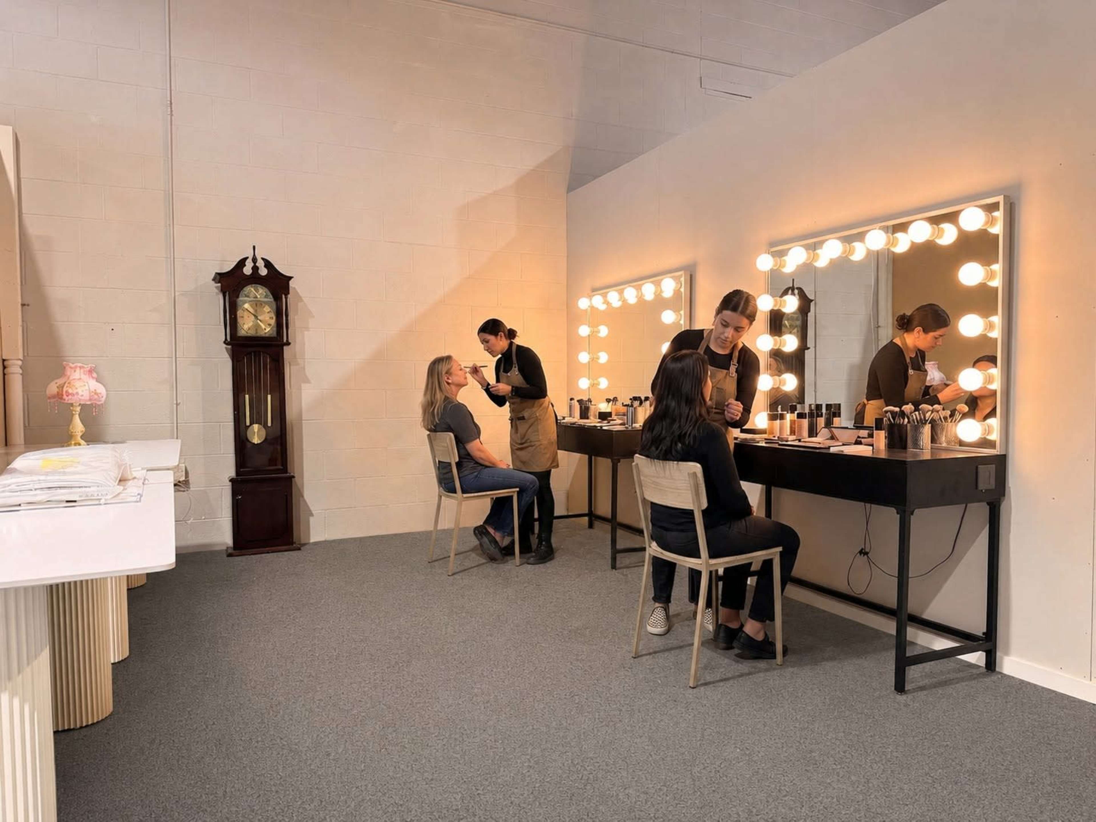 Two clients sit in chairs while makeup artists apply makeup at a well-lit vanity station with large mirrors and bright lights in a modern studio setting.
