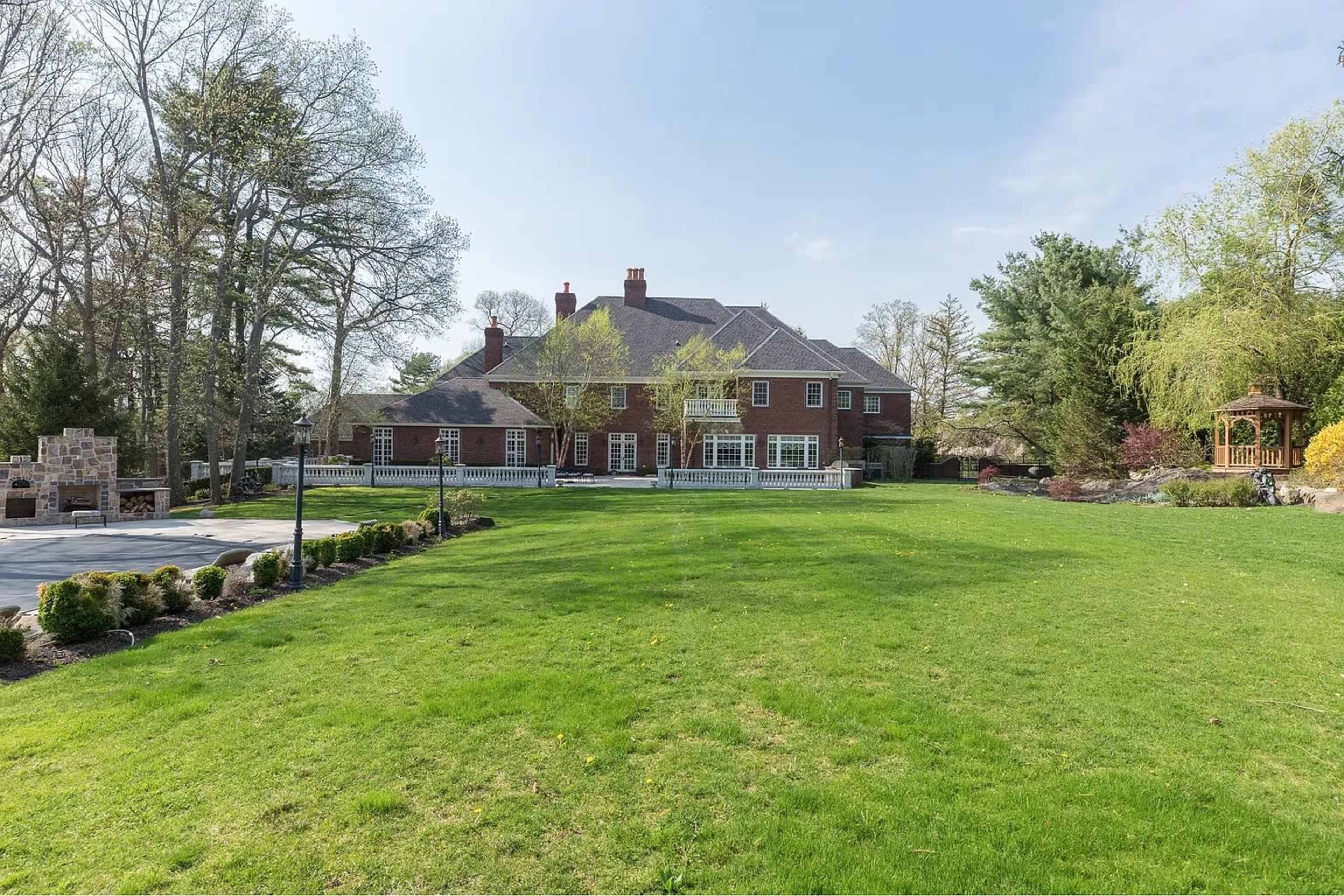 A large brick mansion is situated on a green lawn surrounded by trees and landscaped gardens.