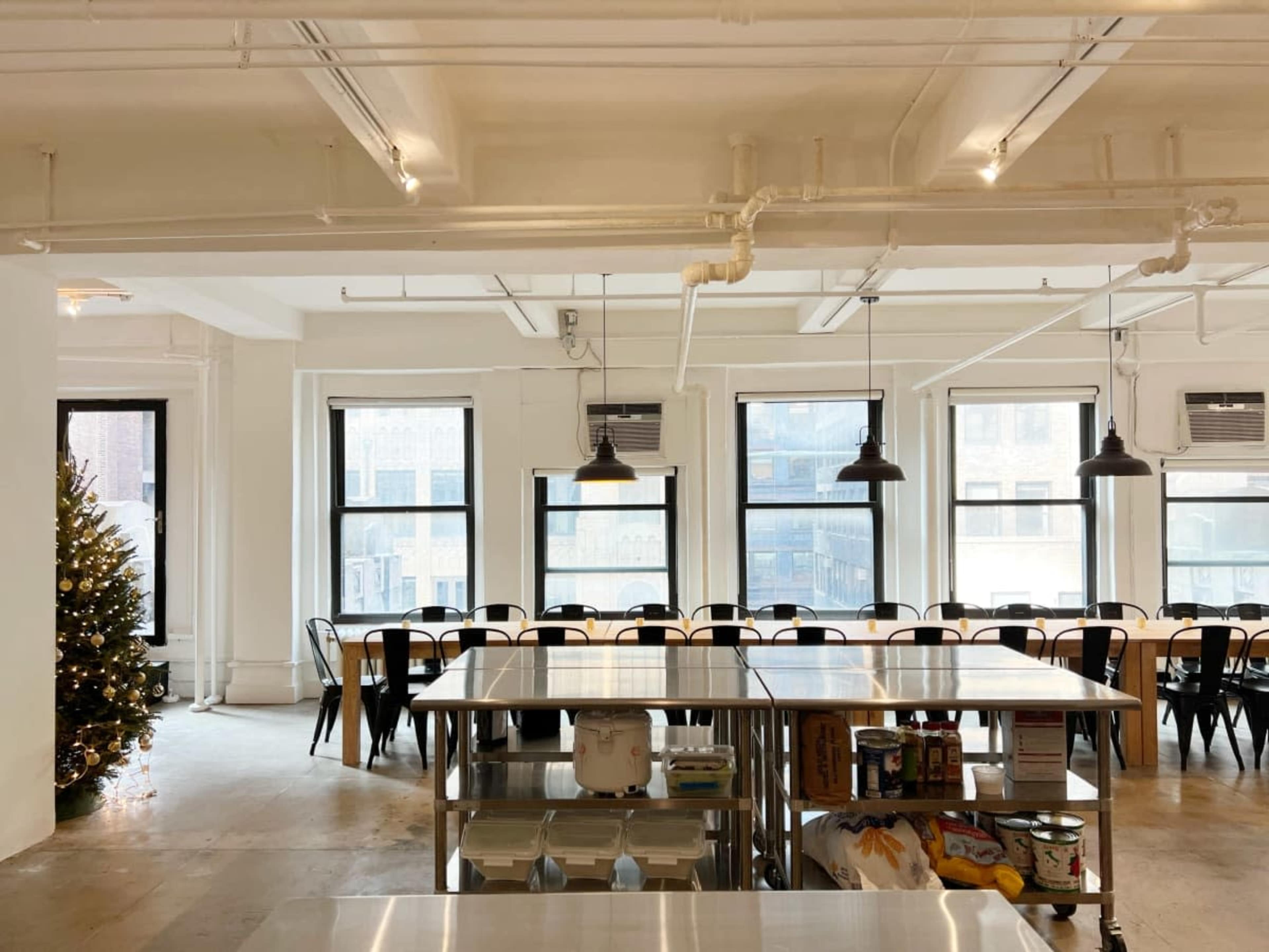 The image shows a bright, open room with large windows, a row of black chairs and tables, and a Christmas tree in one corner.