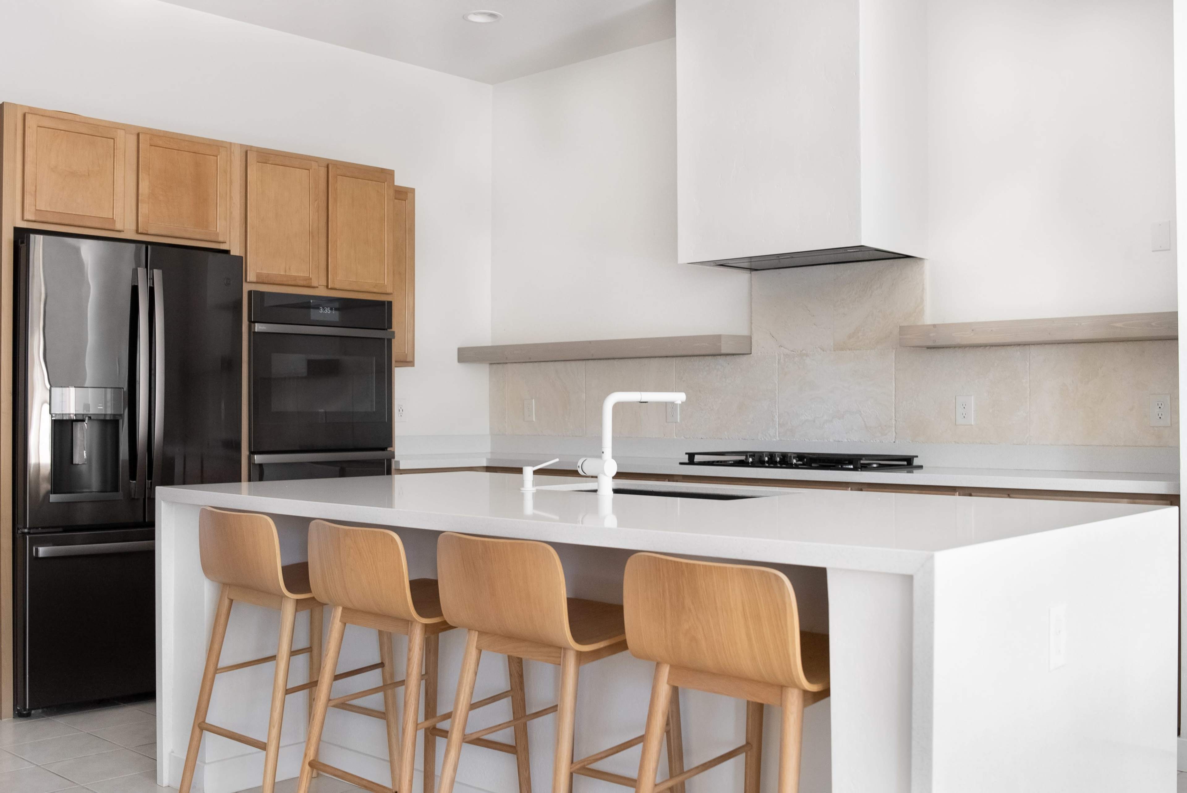 A modern kitchen with wooden cabinets, stainless steel appliances, and a white countertop with bar stools.