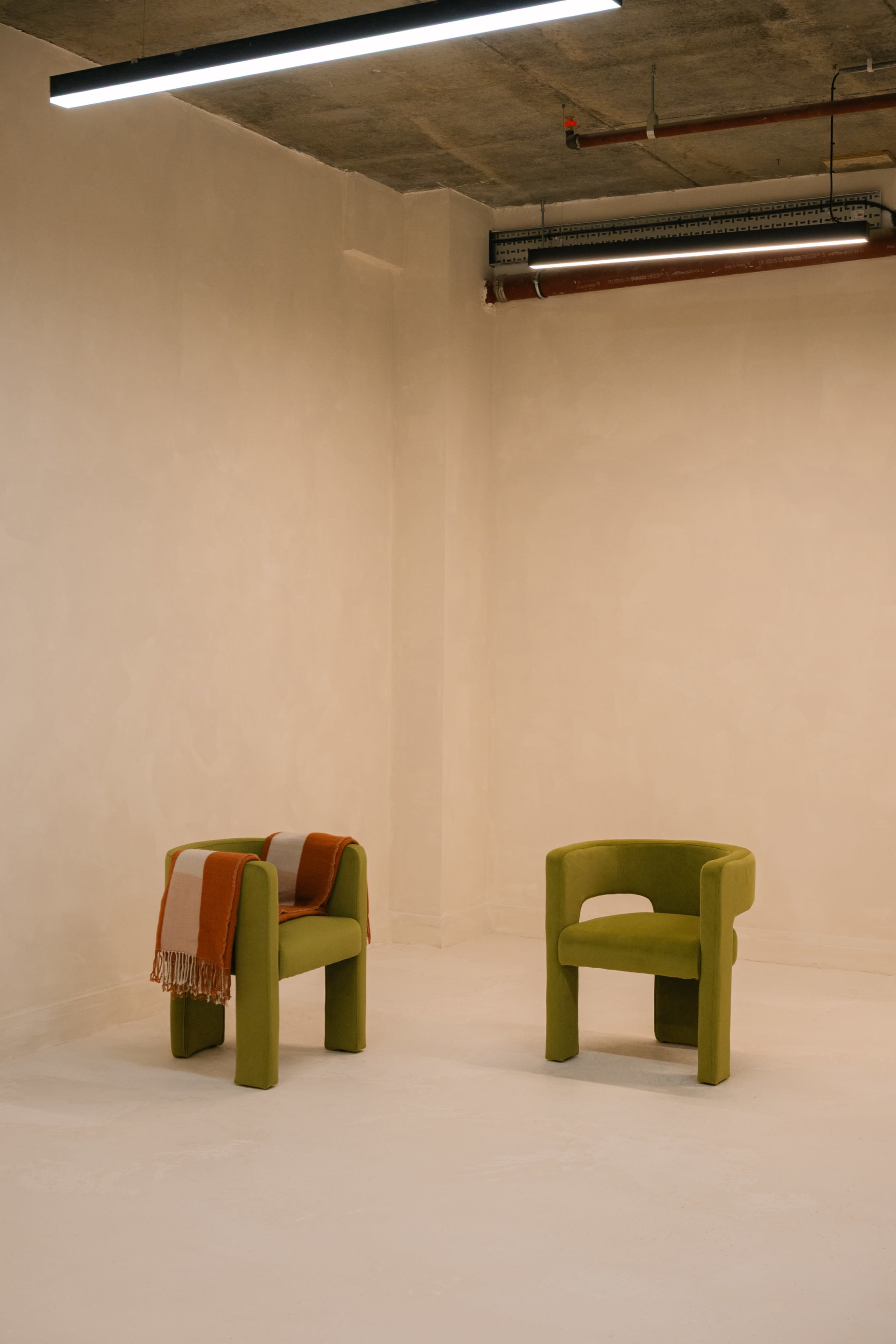 Two green chairs with a throw draped over one are positioned in a minimalistic, empty room with a concrete ceiling and soft lighting.