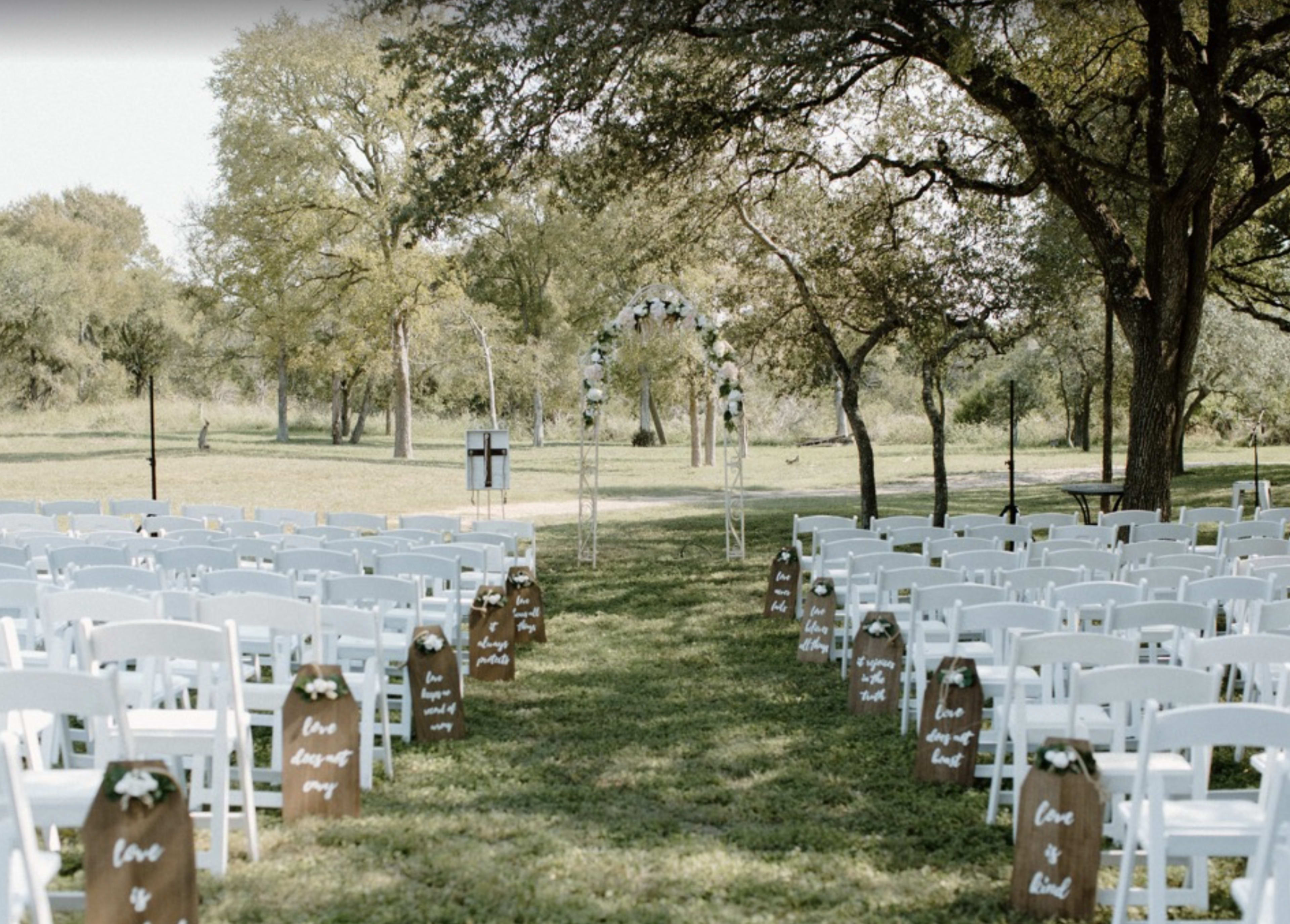 An outdoor wedding ceremony setup features rows of white chairs lining a grassy aisle leading to an arbor decorated with greenery and flowers.