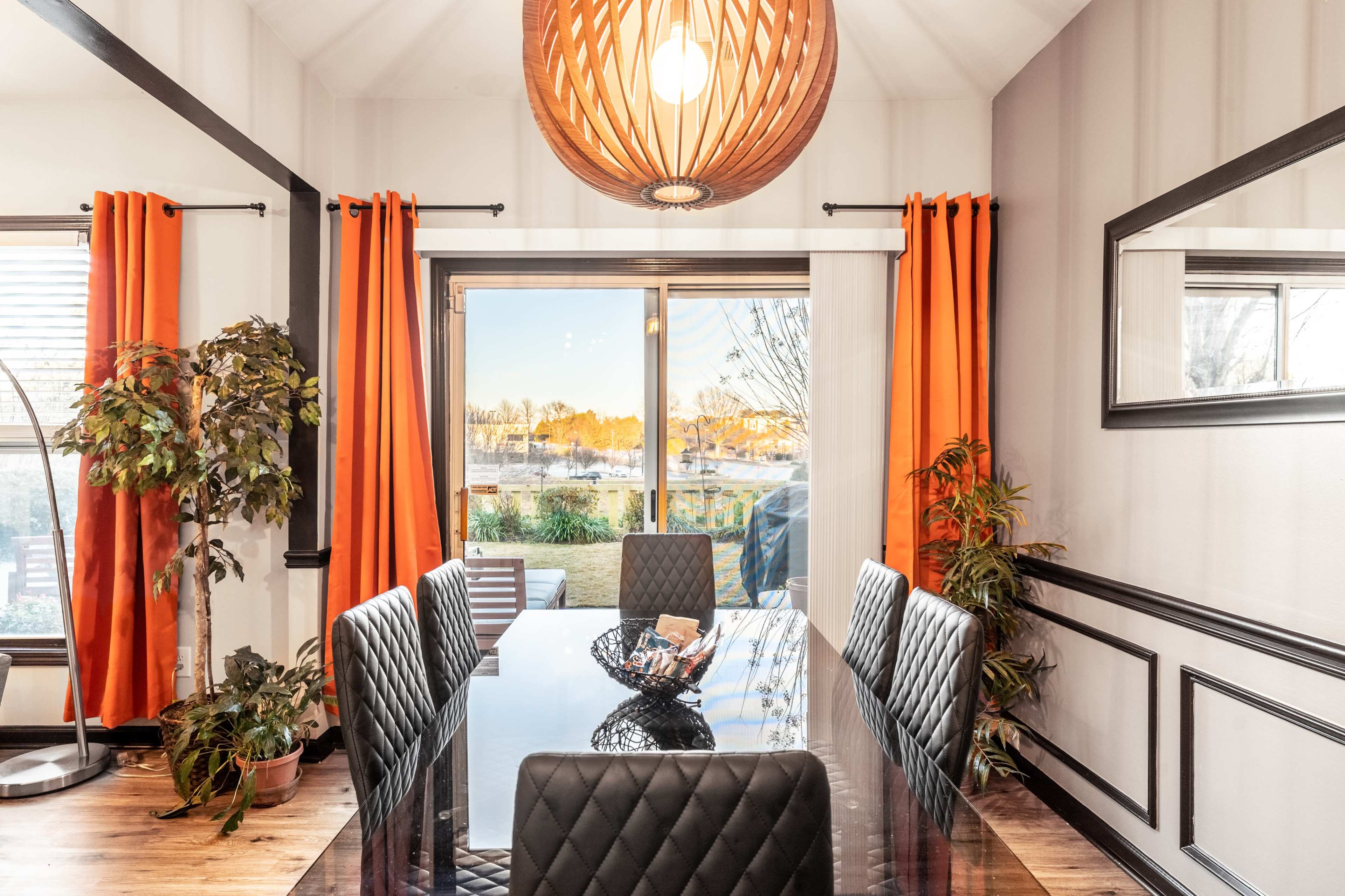 A dining room features a large black table surrounded by six black chairs, orange curtains framing a window that overlooks a patio, and a decorative pendant light hanging from the ceiling.