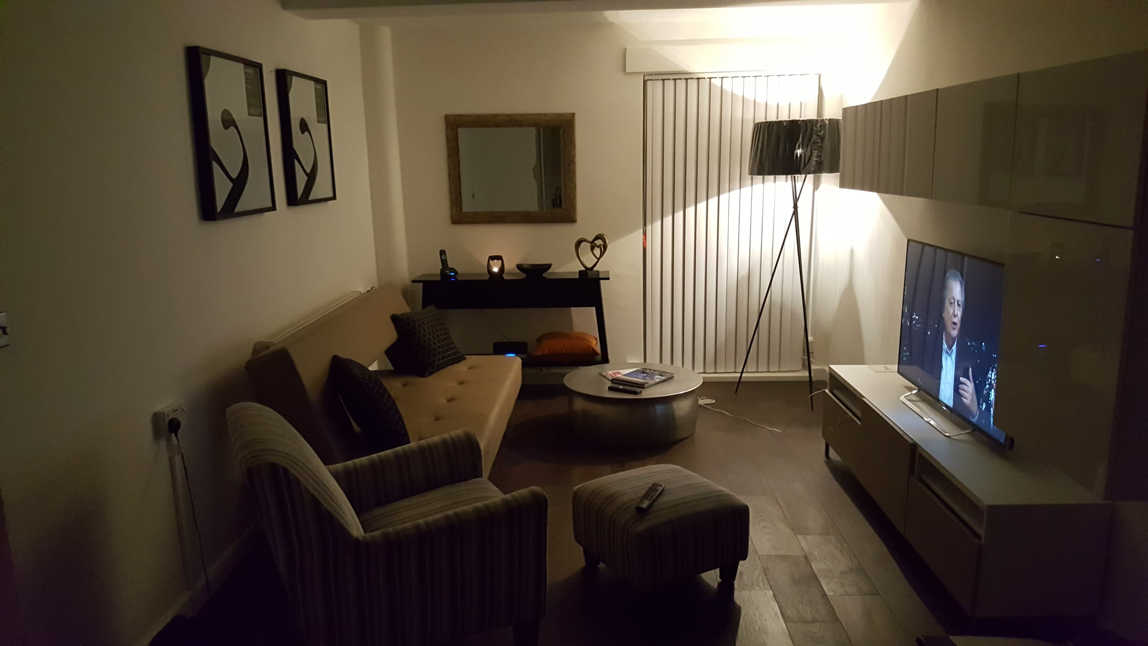A living room with a sofa, small ottoman, television, and a side table, illuminated by a lamp in the corner.