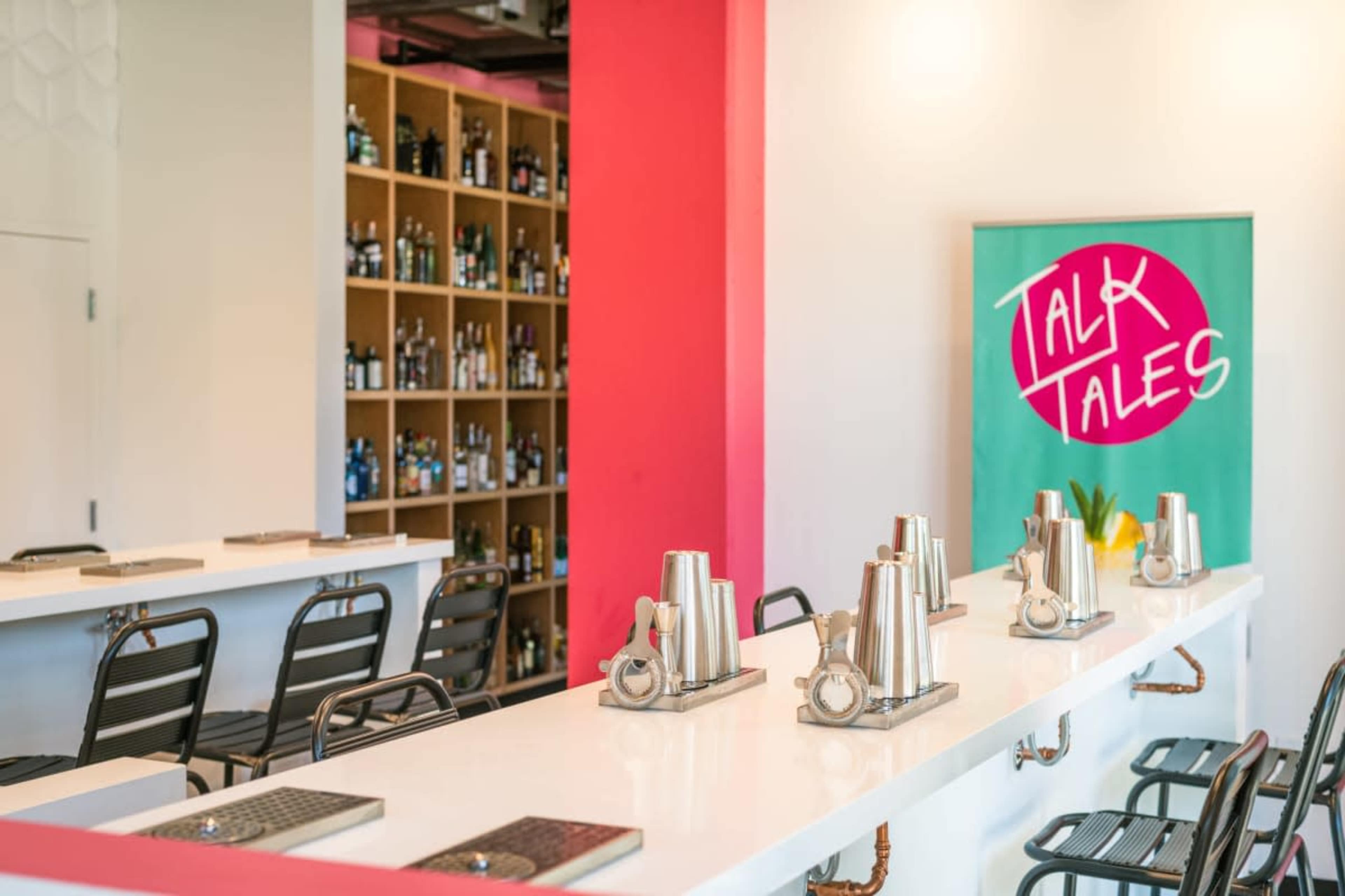The image shows a sleek bar area with a white countertop, metal shaker sets lined up, and a colorful backdrop featuring shelves filled with various bottles.