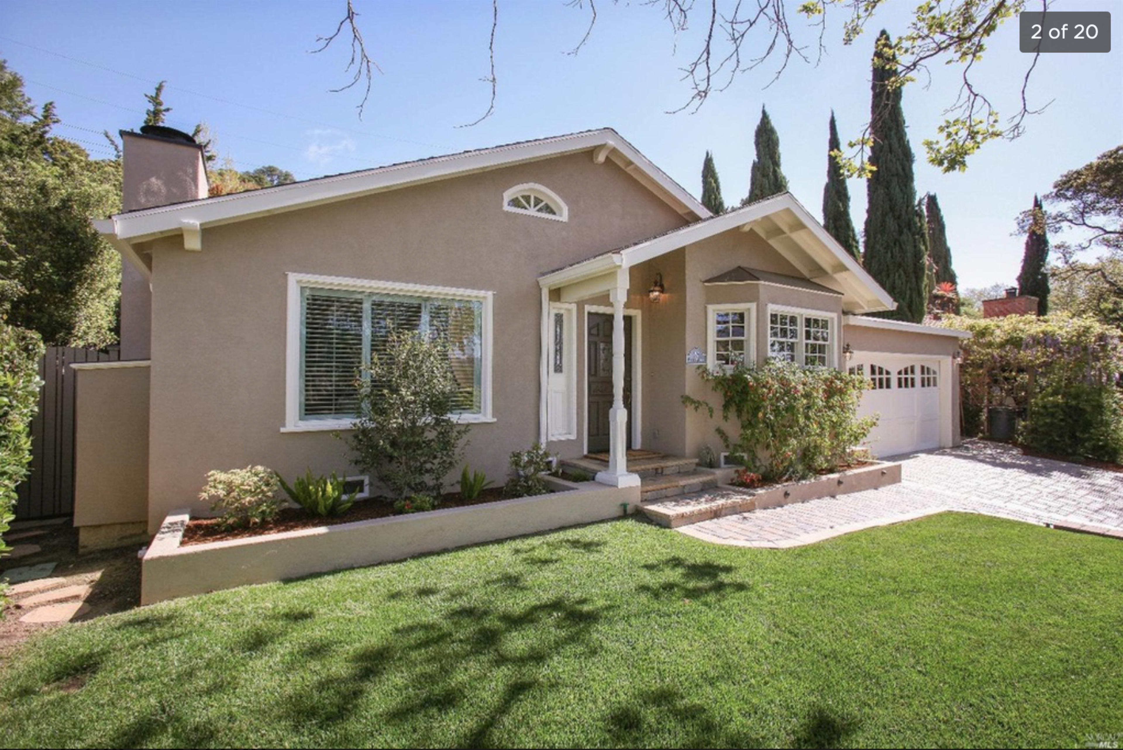 A single-story house with a light beige exterior, a small front porch, and a well-maintained lawn surrounded by greenery.