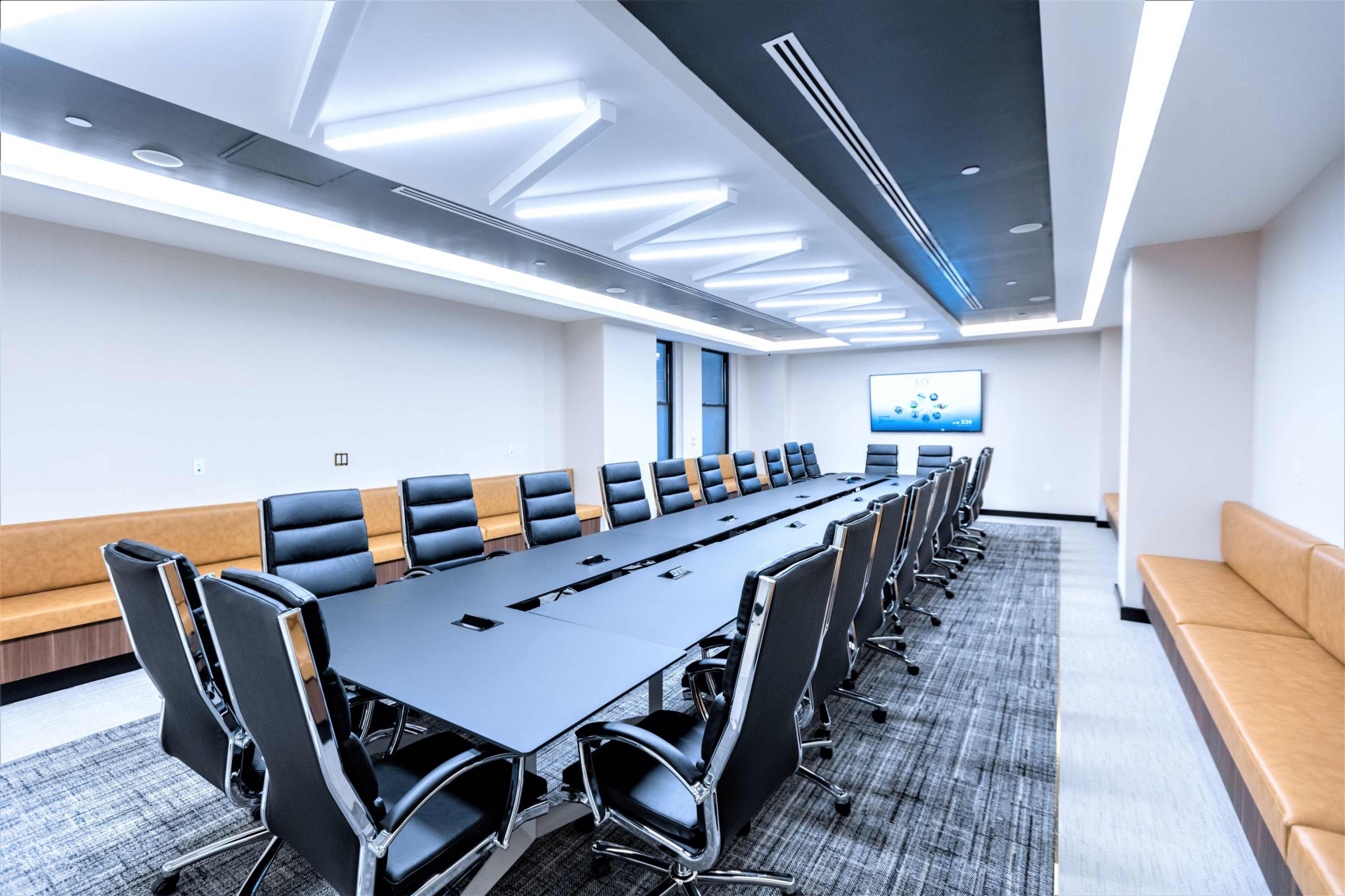 A modern conference room features a long black table surrounded by black leather chairs, with a large screen mounted on the wall.