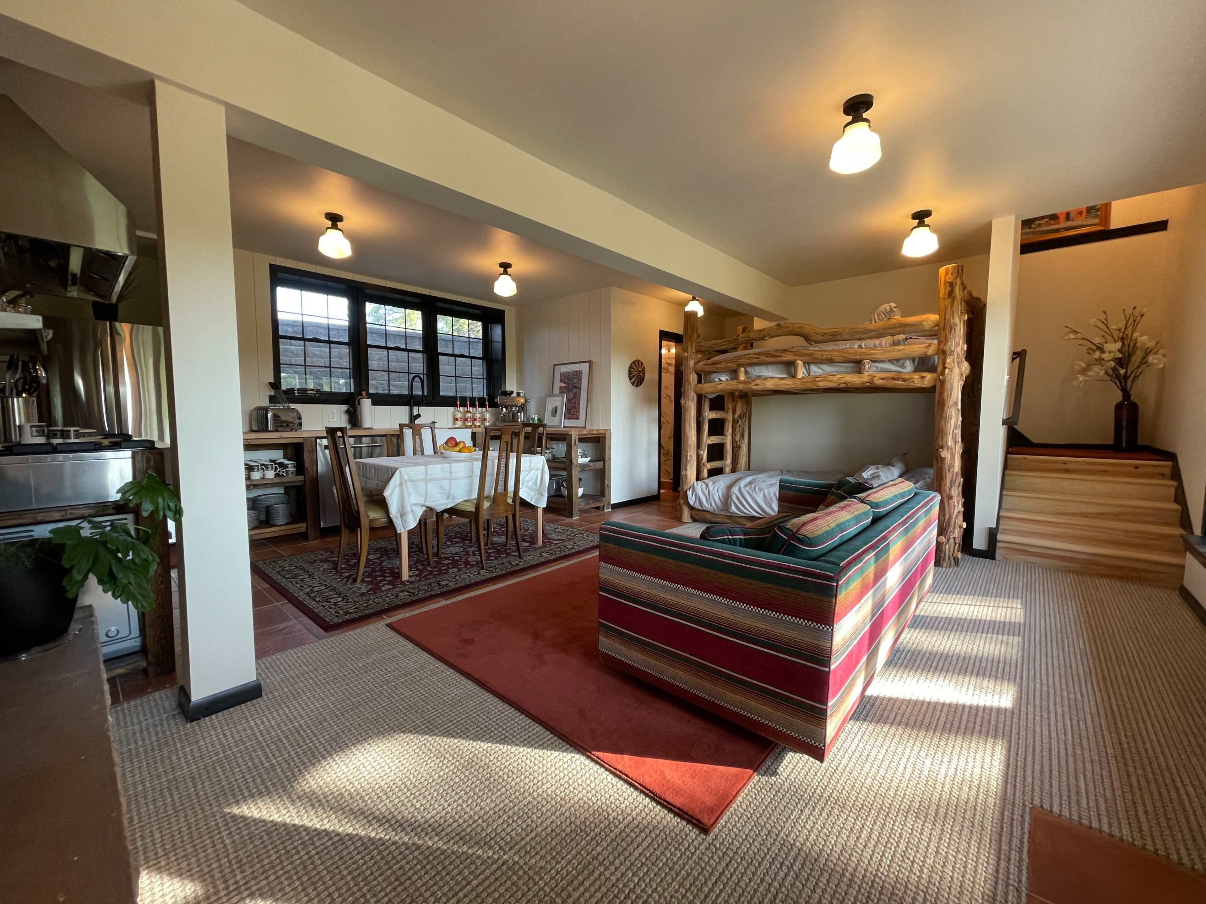 A spacious living area with a striped sofa, a wooden bunk bed, a dining table set for four, and a staircase leading to an upper level.