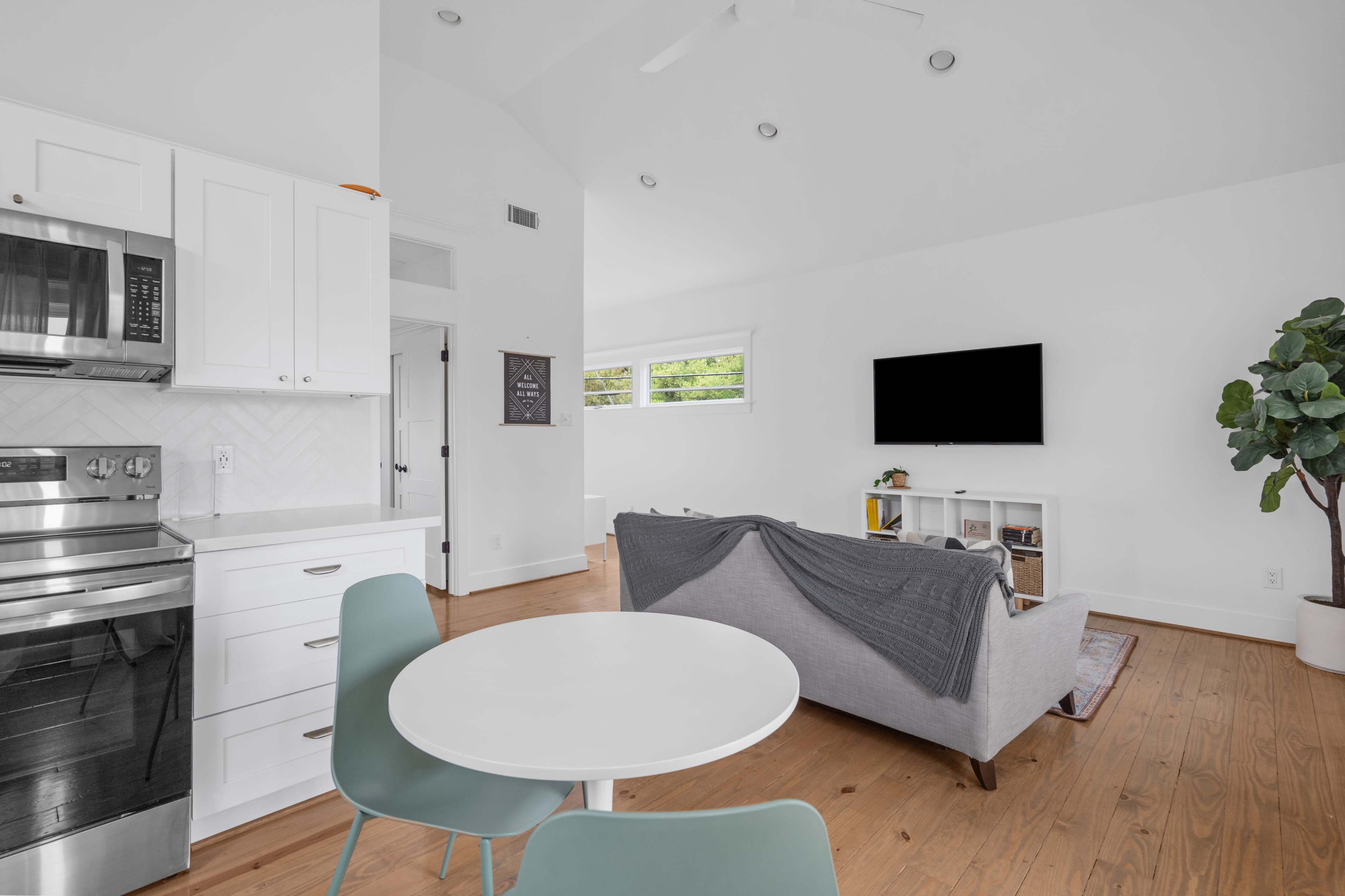 The image shows a modern, open-concept living space featuring a kitchen with white cabinetry, a dining table, and a living area with a sofa and a television on the wall.