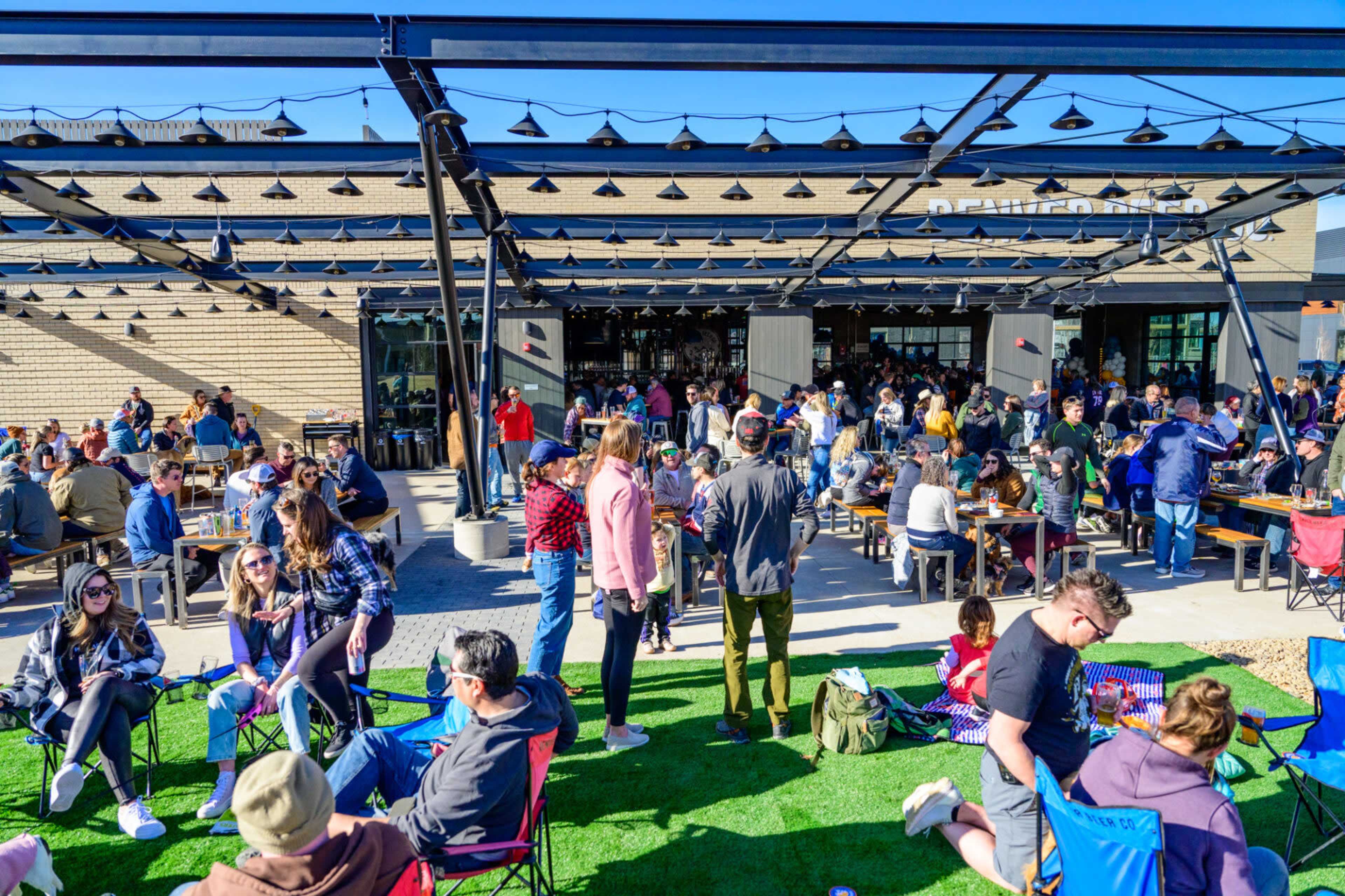 A large outdoor gathering is taking place at a brewery, with many people seated at tables and some in lawn chairs on a grassy area.