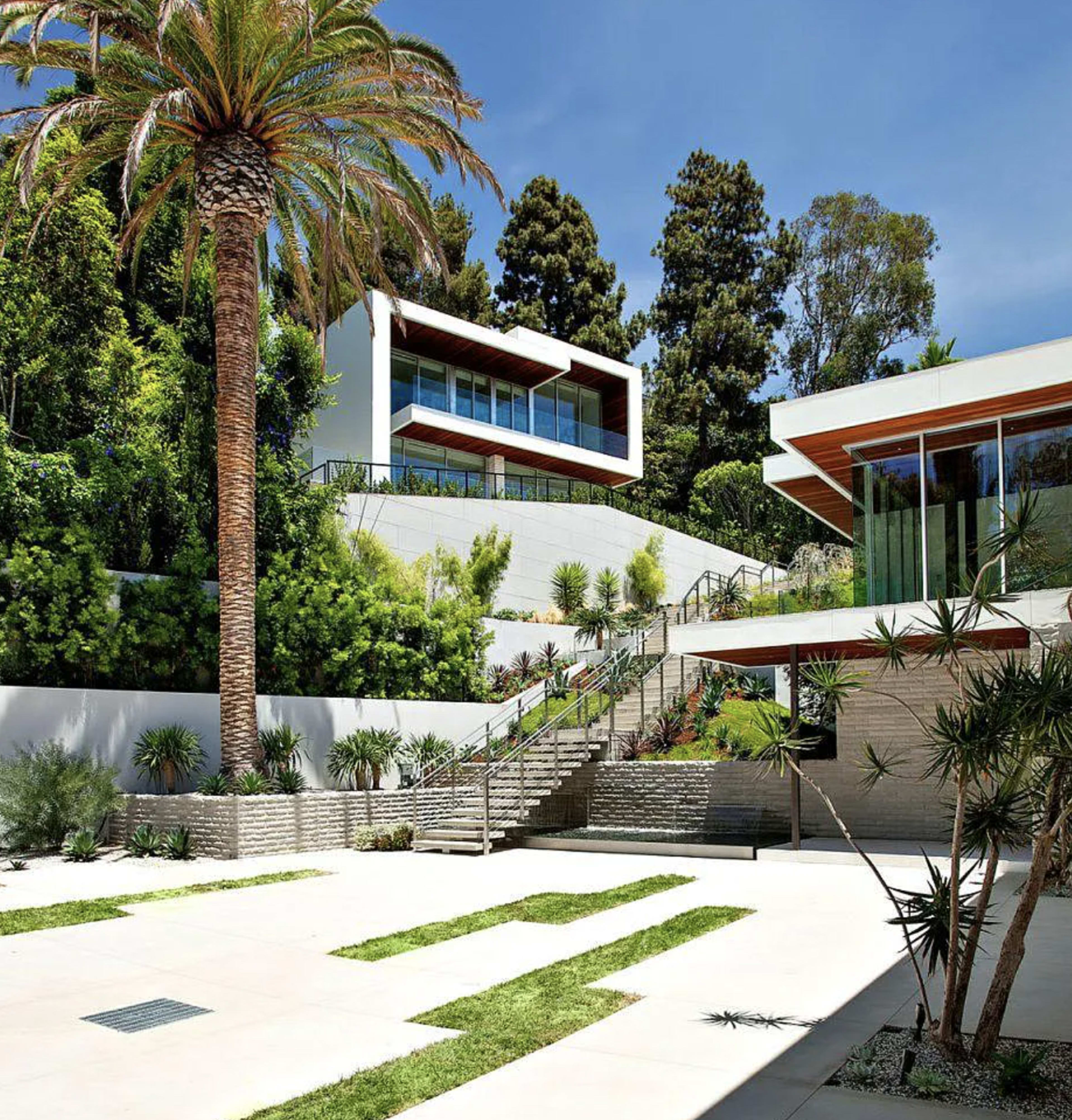 A modern house with large windows is set on a landscaped hillside, featuring palm trees and stone steps leading up to the entrance.