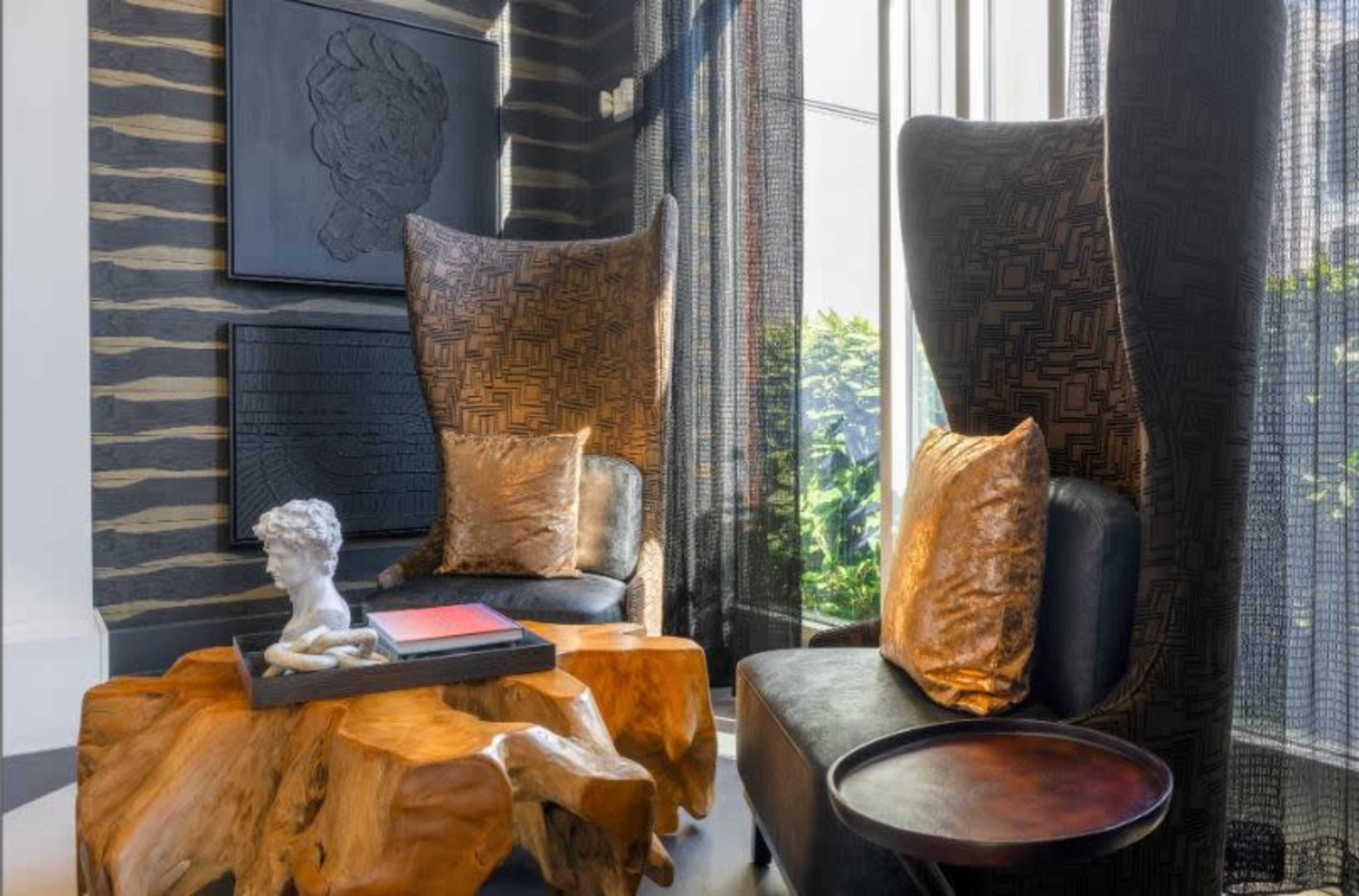 The image shows a stylish seating area featuring two tall-backed chairs, a unique wooden coffee table, and decorative art against a patterned wall.