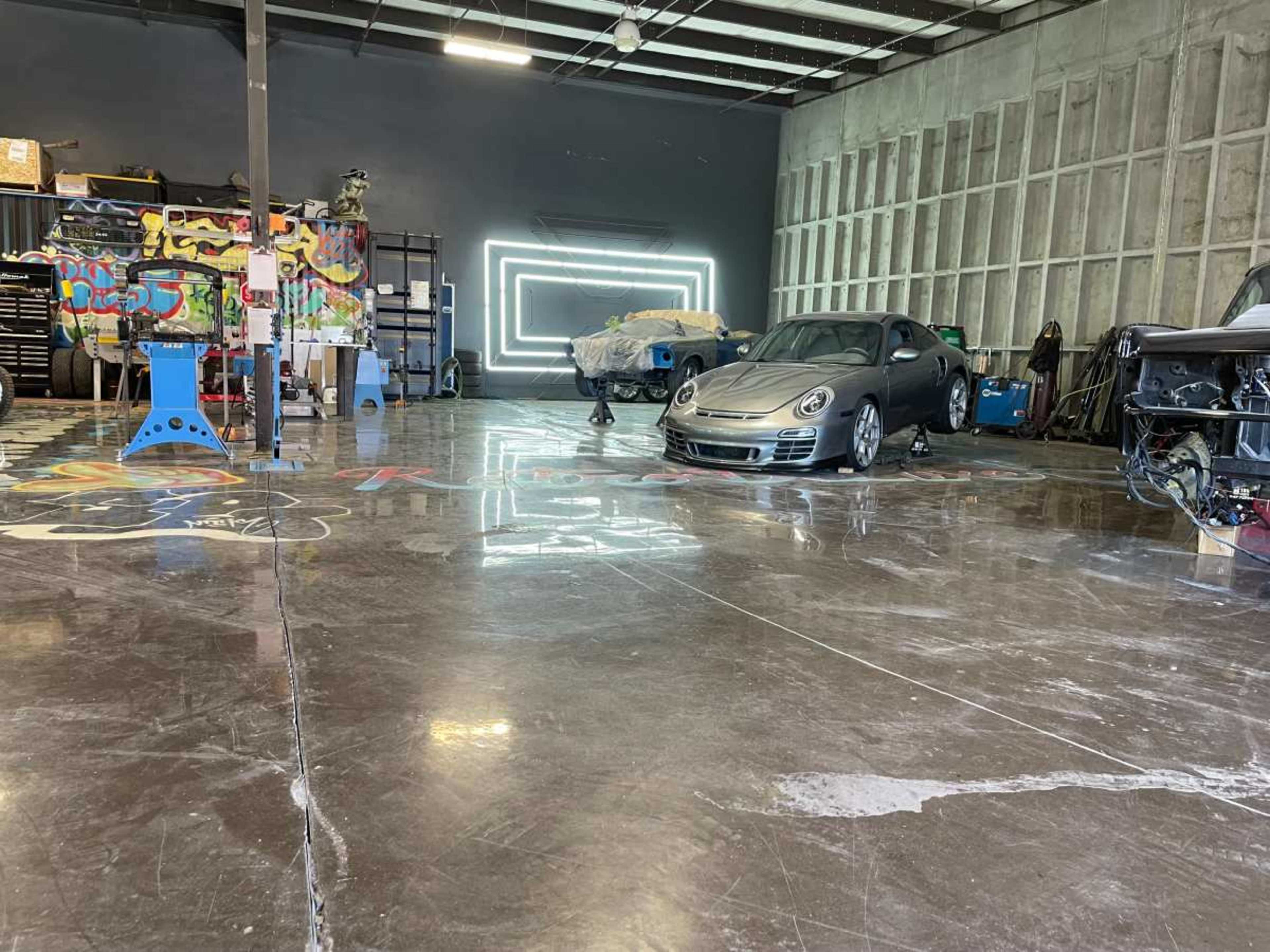 The image shows a spacious automotive workshop with polished concrete floors, featuring a gray car in the foreground and various tools and equipment in the background.