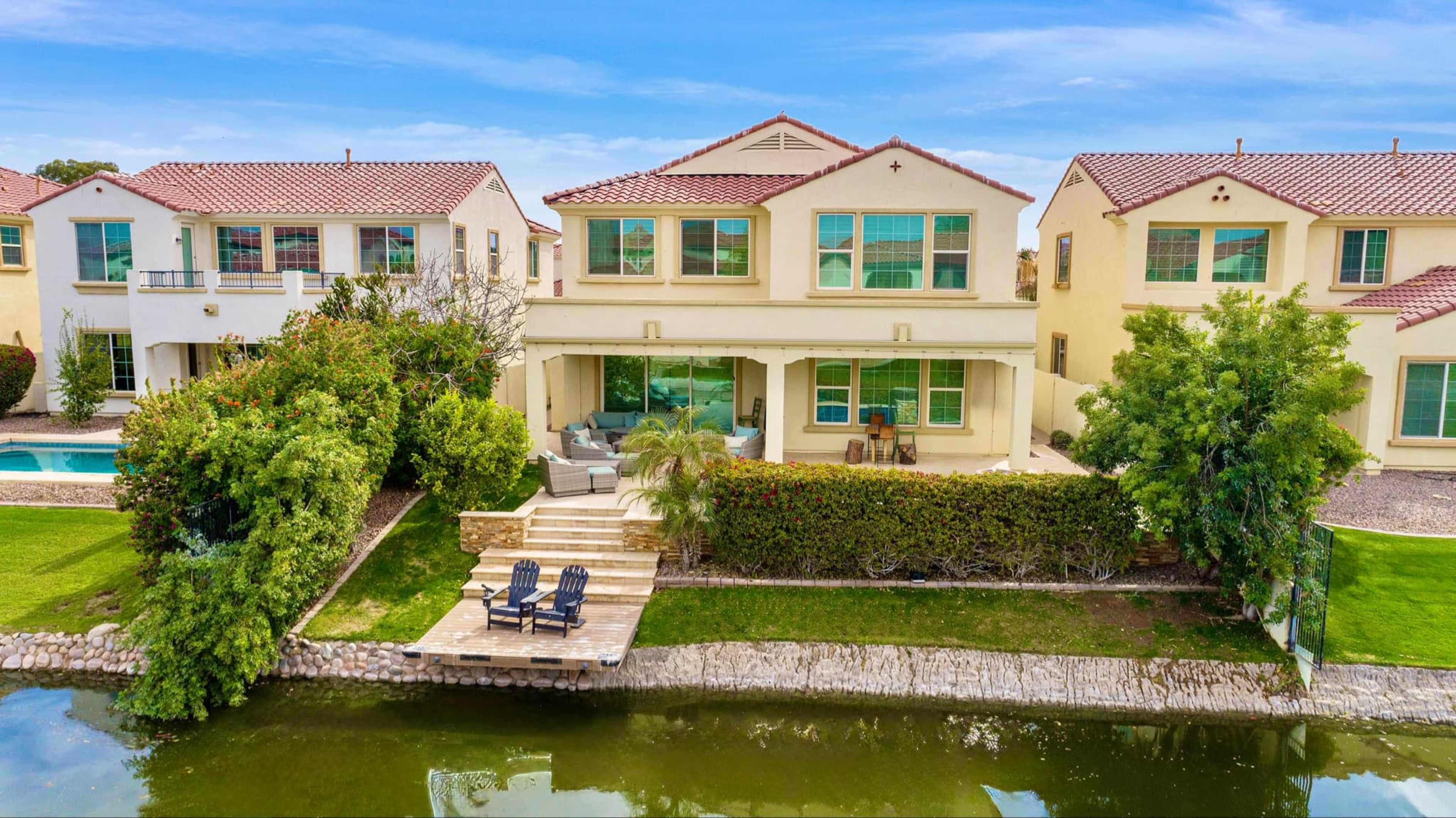 A two-story house with a backyard patio overlooks a small body of water, flanked by neighboring homes and landscaped greenery.