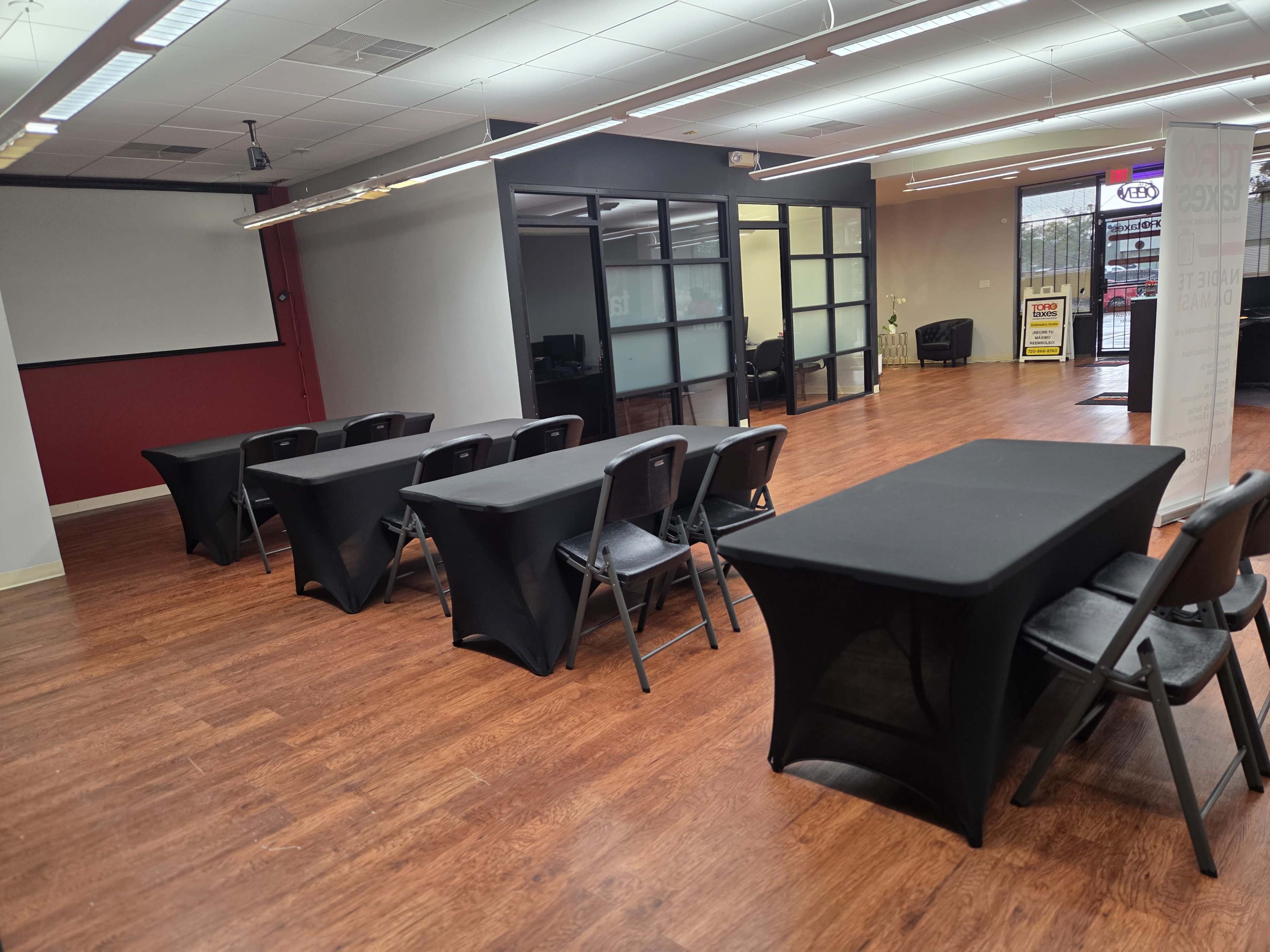 The image shows a spacious room with several black table setups and a glass-walled office area in the background.
