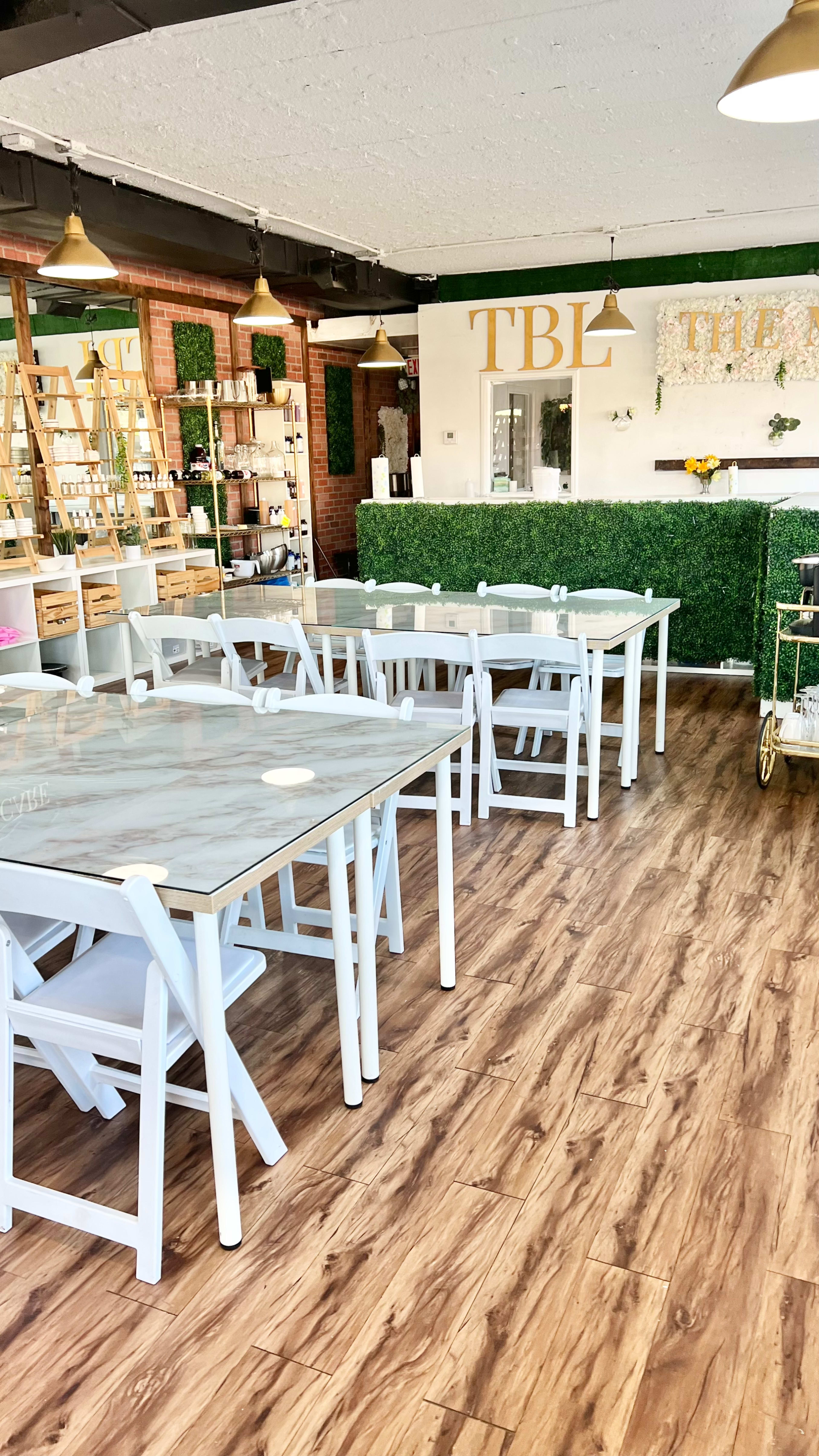 A bright, modern café with white tables and chairs arranged on a wood-patterned floor, featuring green wall decorations and a service counter in the background.
