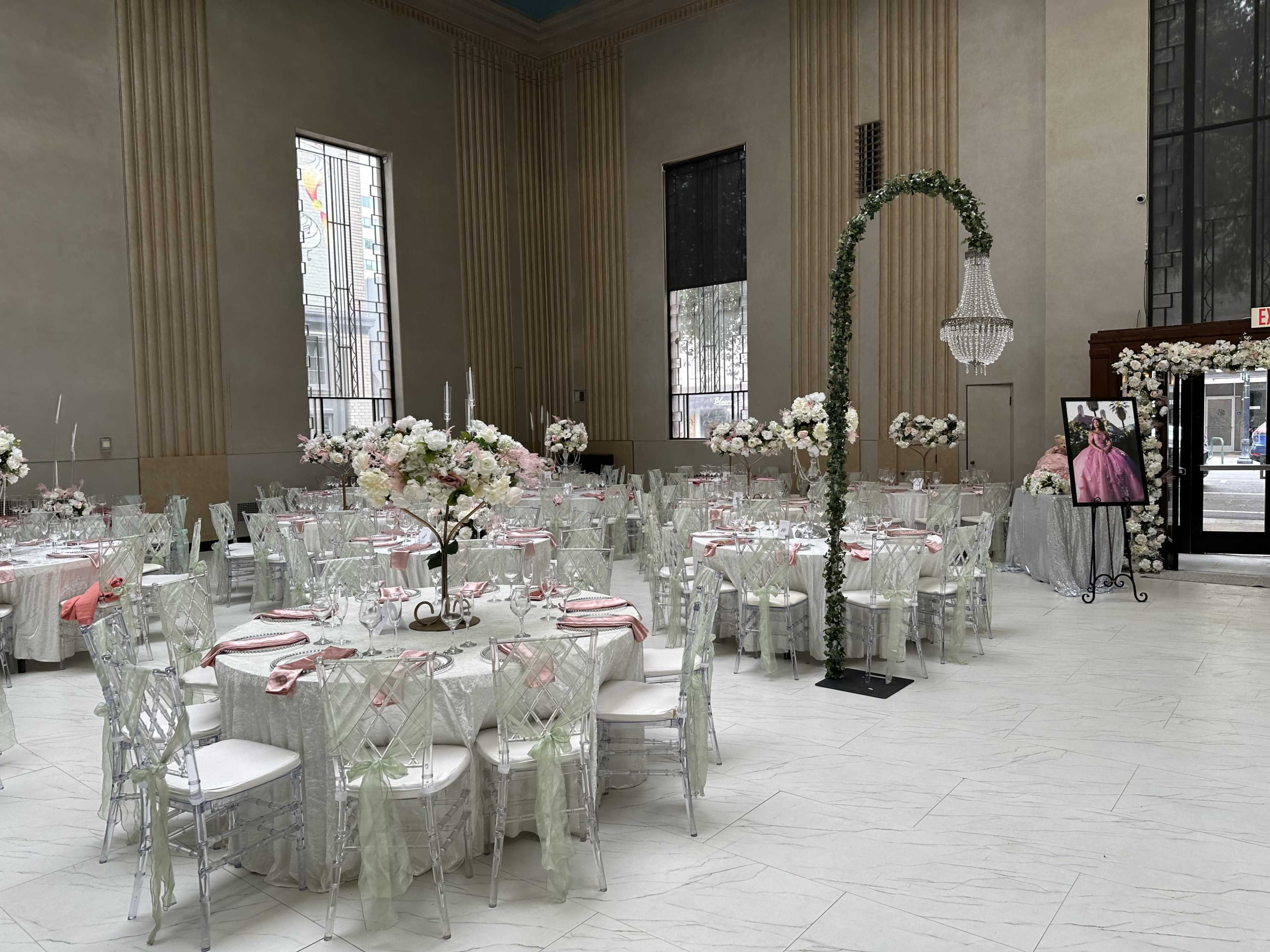 The image shows an elegantly decorated banquet hall with round tables set for a formal event, featuring floral centerpieces and ornate chairs.