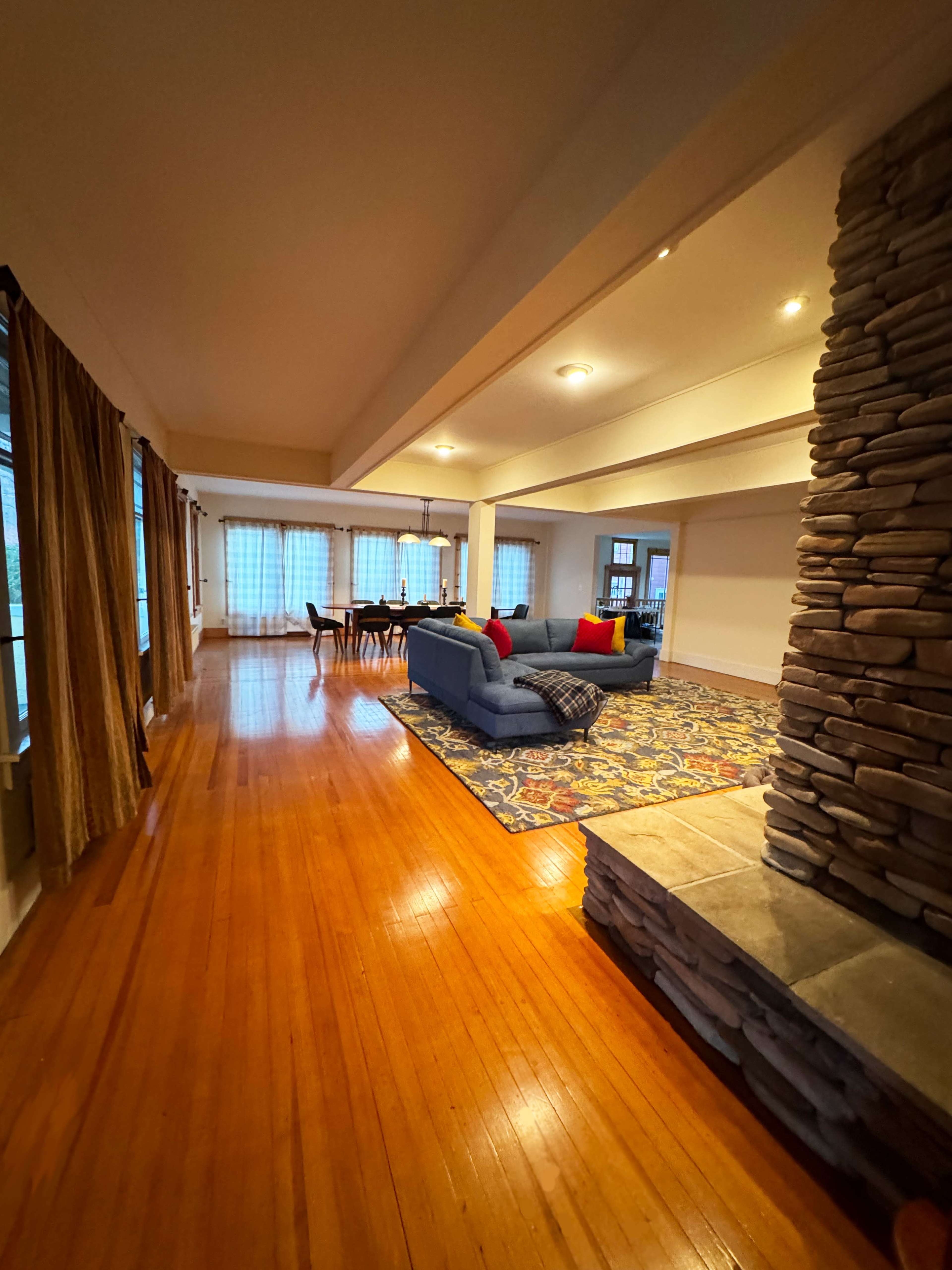 The image shows a spacious living area with wooden flooring, a stone fireplace, a blue sofa adorned with colorful pillows, and a dining table in the background.