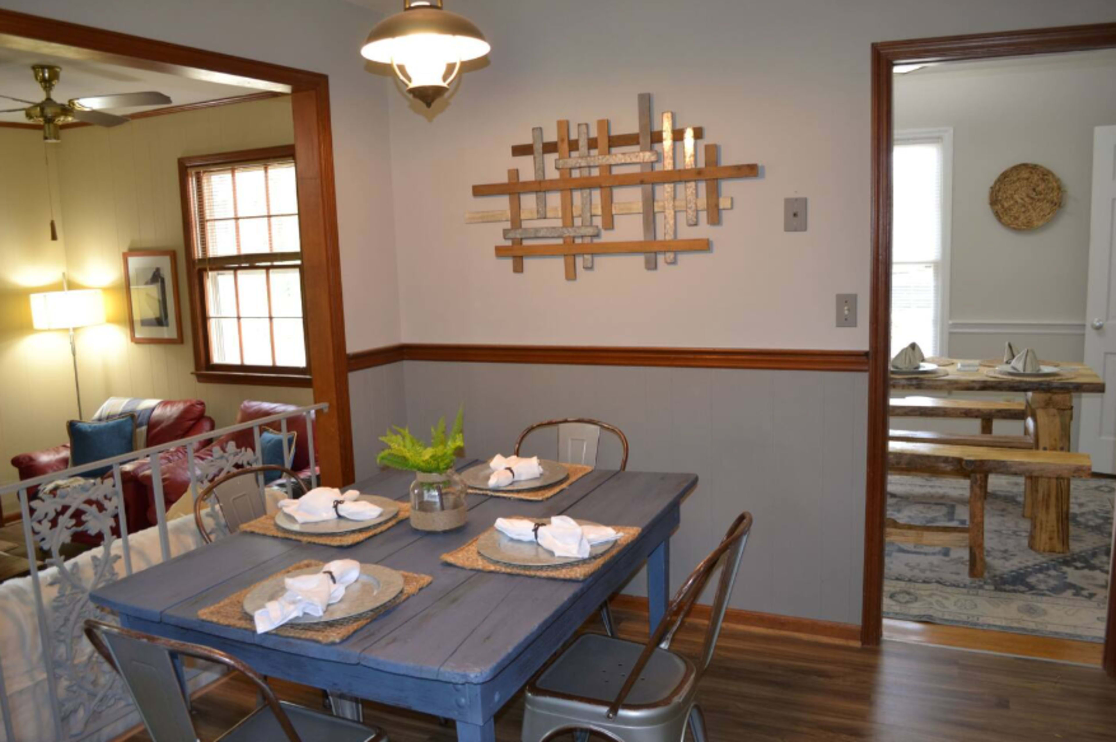 A dining area features a blue table set with white napkins and plates, surrounded by metal chairs, and is adjacent to a living space with a red couch visible through an open doorway.