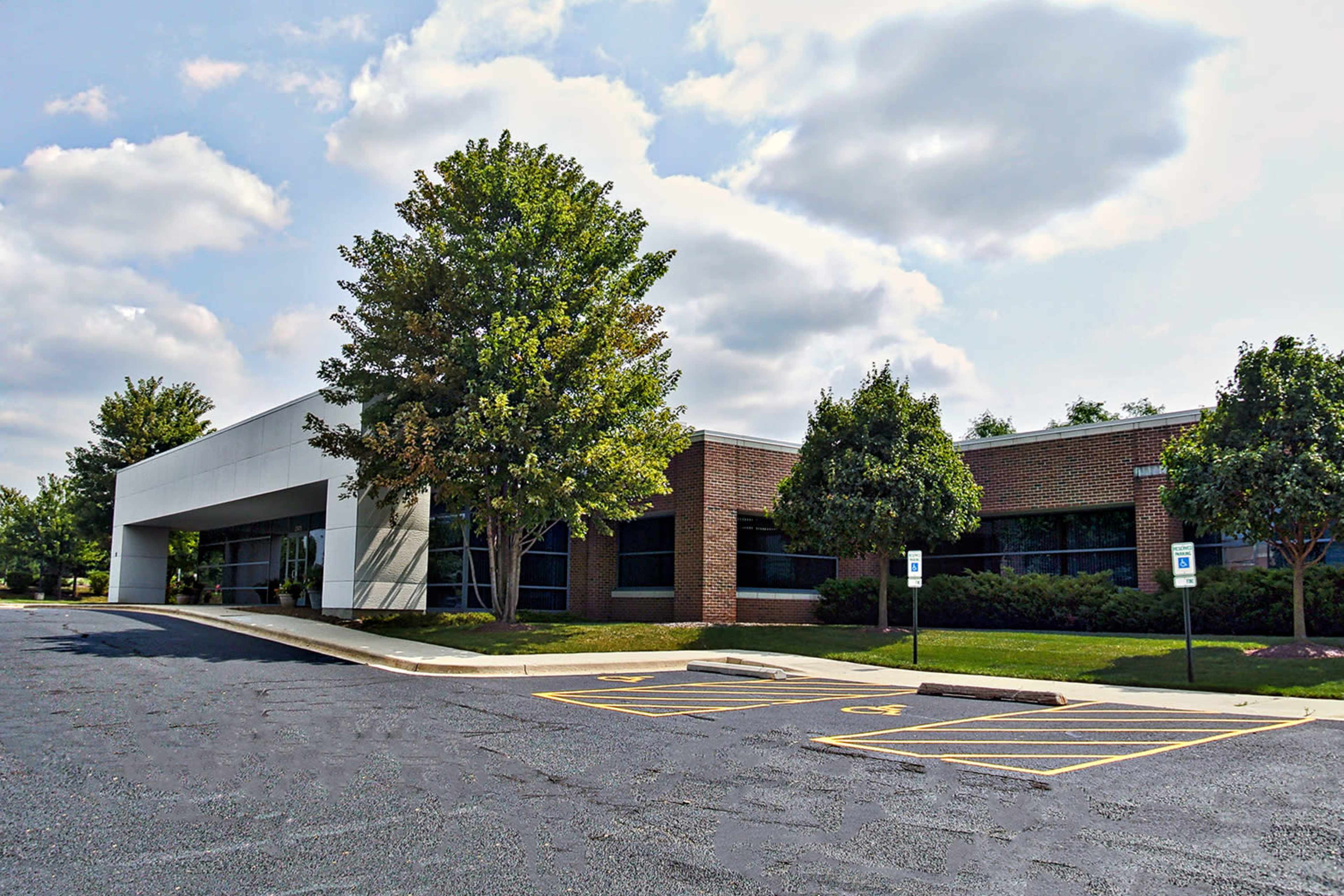 The image shows a modern commercial building with a landscaped parking area featuring several trees and a clear sky.