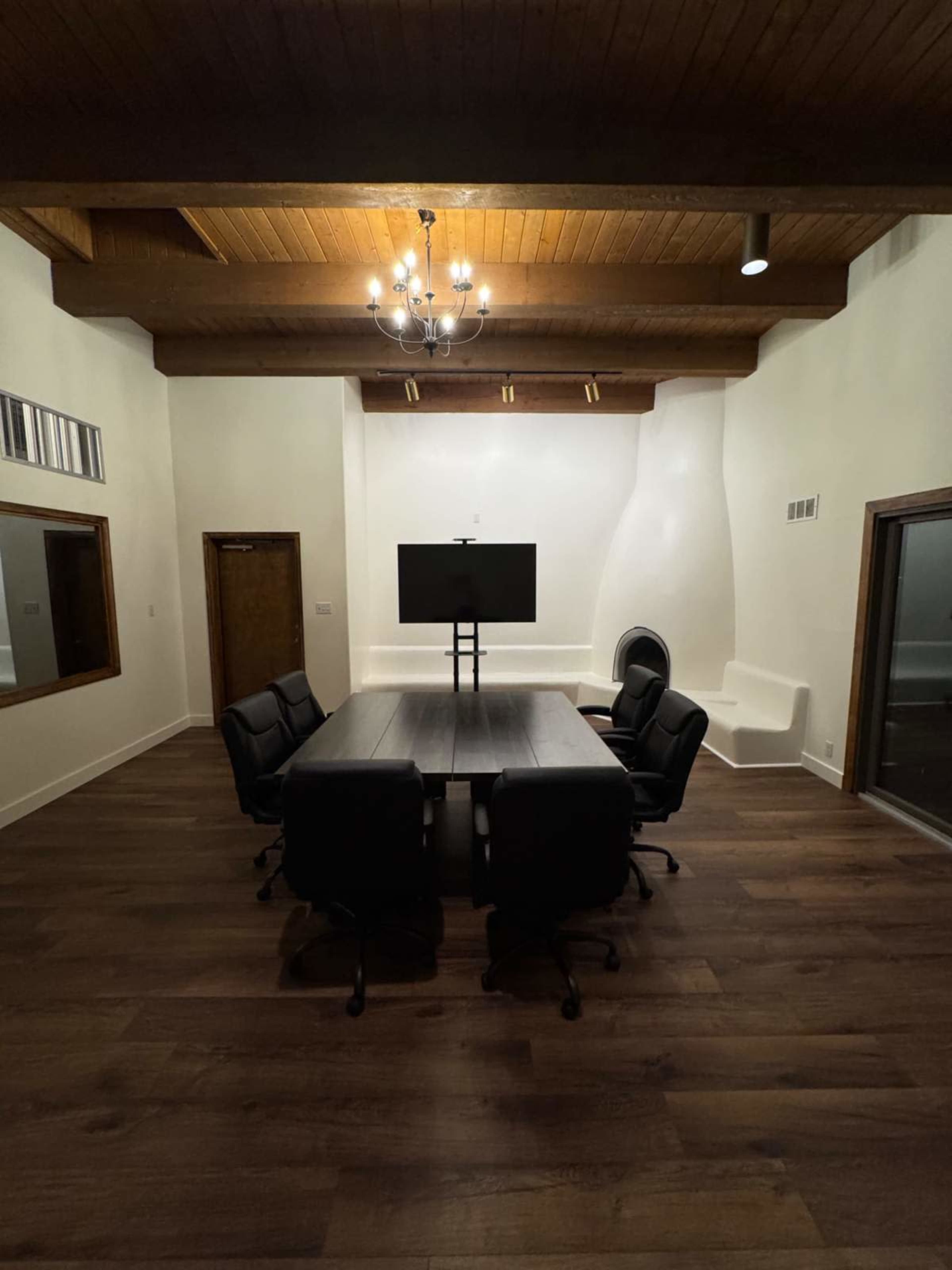 A conference room features a large wooden table surrounded by six black chairs, with a television mounted on the wall and a chandelier above.