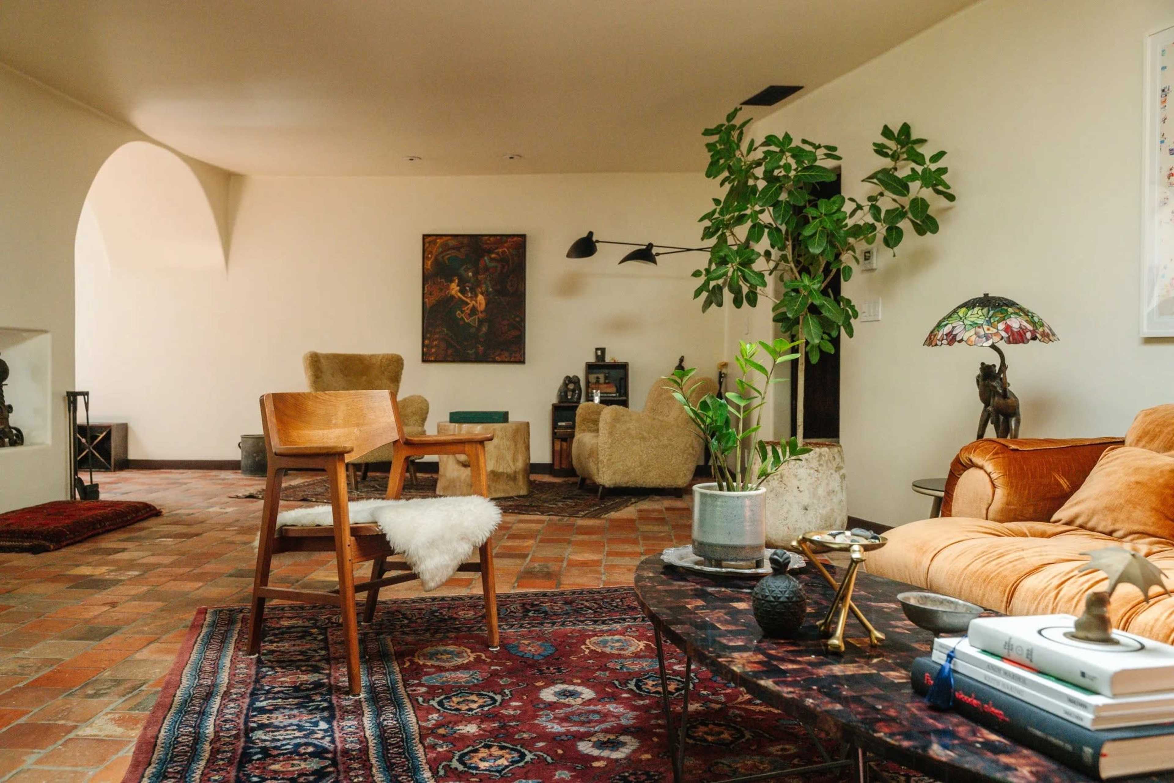 The image shows a spacious living area with terracotta tiled flooring, a mix of comfortable seating, a decorative table, and green plants.